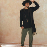 A man in a wide-brimmed hat and black shirt, accessorized with a long necklace and scarf, stands barefoot by a pool. He wears AYA Sacred Wear's dark green long cotton pants that add an artistic touch against the textured beige wall backdrop.