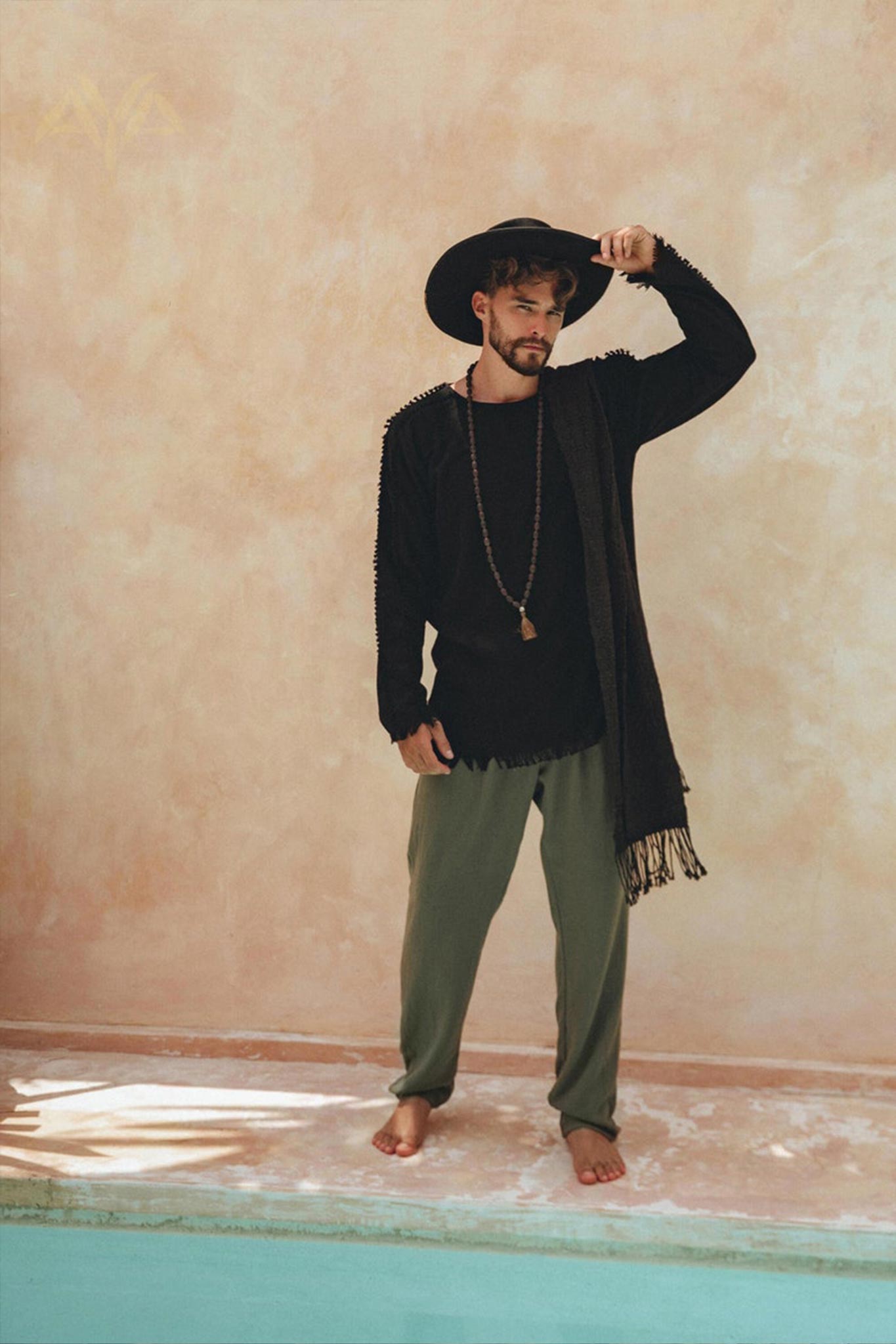 A man in a wide-brimmed hat and black shirt, accessorized with a long necklace and scarf, stands barefoot by a pool. He wears AYA Sacred Wear's dark green long cotton pants that add an artistic touch against the textured beige wall backdrop.