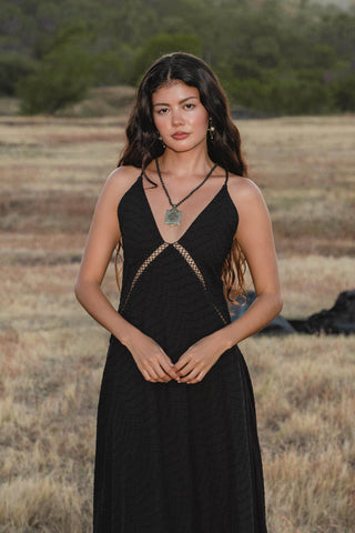 A woman with long wavy brown hair stands in a dry grassy field, calmly facing the camera and wearing the AYA Sacred Wear Dhara Loom Dress in Black—a handwoven cotton boho bridesmaid dress with geometric cutouts. Green hills rise behind her.