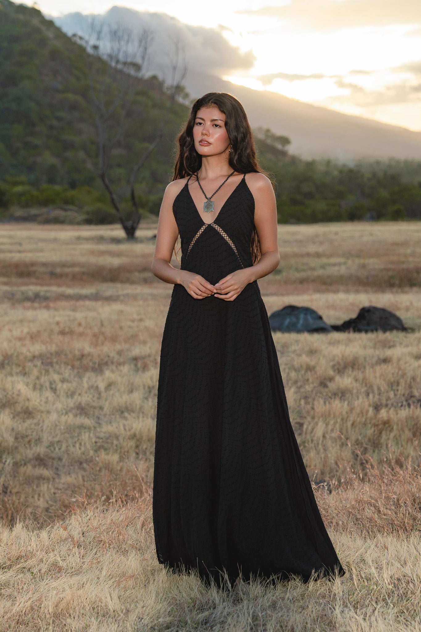 A woman with long, wavy hair stands in a dry grassy field at sunset, wearing the AYA Sacred Wear Dhara Loom Dress in Black—a handwoven cotton boho bridesmaid dress with a V-neck, cutout detail, and handcrafted knot embroidery. Misty mountains rise behind her.