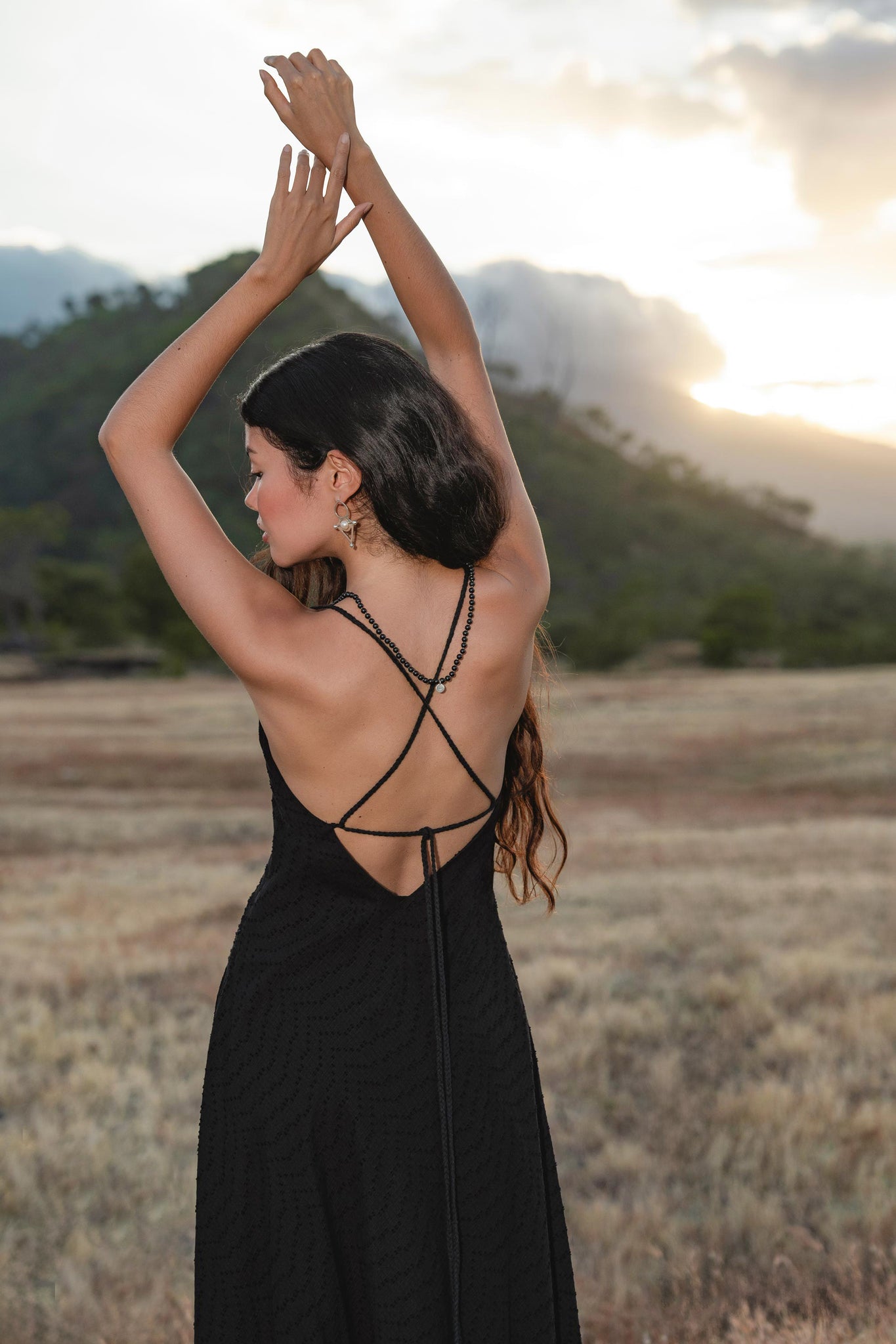 A woman stands in a field at sunset, arms raised, wearing the AYA Sacred Wear Dhara Loom Dress in black—a handwoven cotton bridesmaid boho dress. Mountains, trees, and clouds form the scenic background.