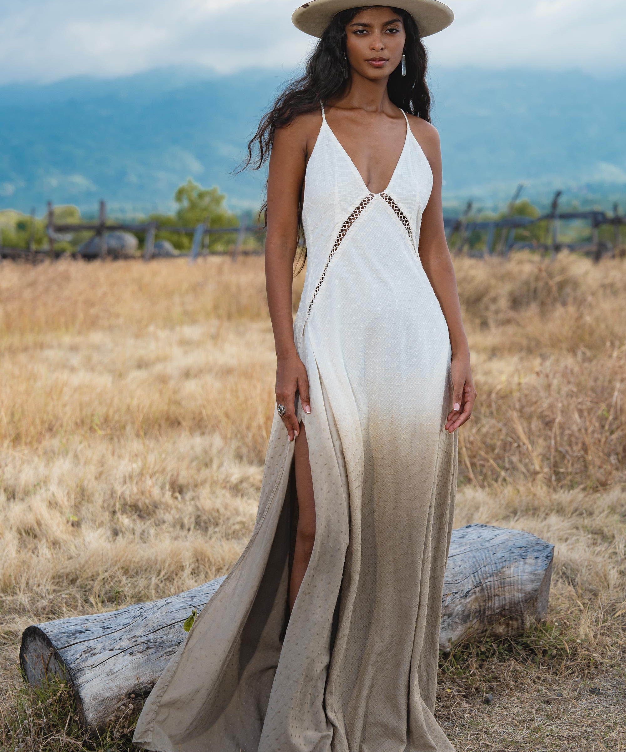 A woman stands on dry grass near a log, wearing the Dhara Loom Dress in Ombre Mocco by AYA Sacred Wear—a backless macrame pure cotton boho dress with a deep V-neck and front slit. Mountains and cloudy sky are in the background.