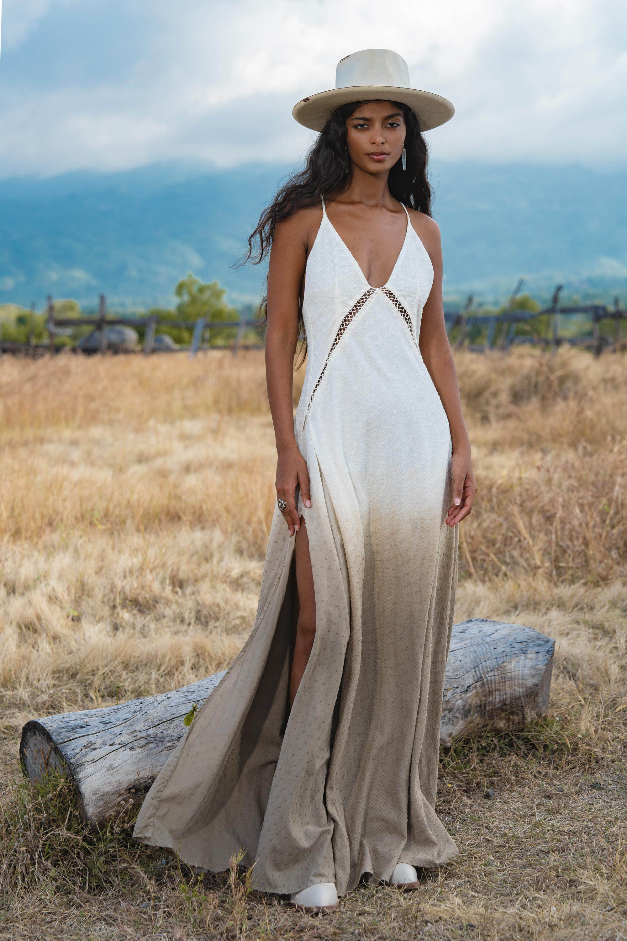 A woman stands on dry grass near a log, wearing the Dhara Loom Dress in Ombre Mocco by AYA Sacred Wear—a backless macrame pure cotton boho dress with a deep V-neck and front slit. Mountains and cloudy sky are in the background.