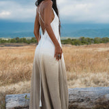 A woman stands in a dry grassy field facing away, wearing AYA Sacred Wear’s Dhara Loom Dress in Ombre Mocco—a backless macrame pure cotton boho dress—with mountains and clouds in the background.
