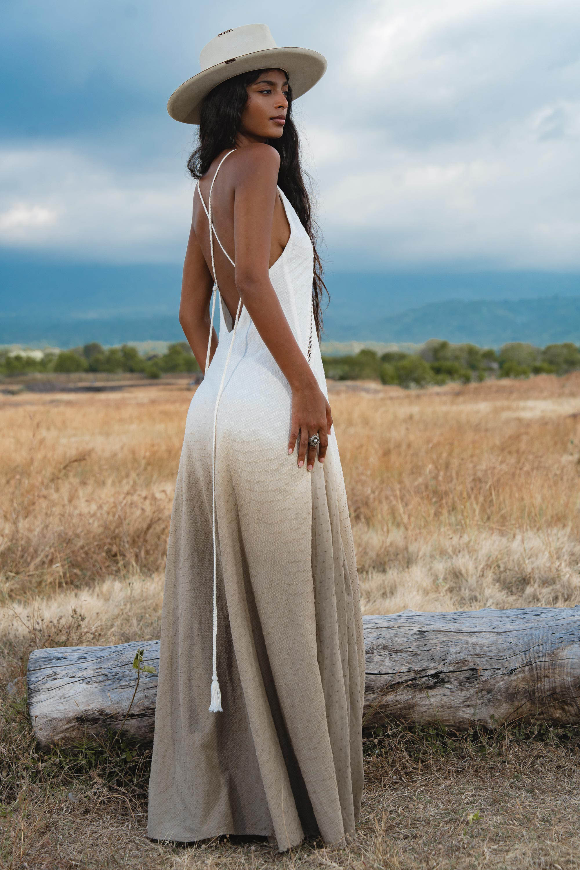 A woman stands in a dry grassy field facing away, wearing AYA Sacred Wear’s Dhara Loom Dress in Ombre Mocco—a backless macrame pure cotton boho dress—with mountains and clouds in the background.