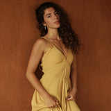 A woman with long curly hair wears the Powder Yellow Padma Dress by AYA Sacred Wear, a sleeveless boho backless dress, standing against a textured brown wall and gazing calmly at the camera while lightly holding her dress.