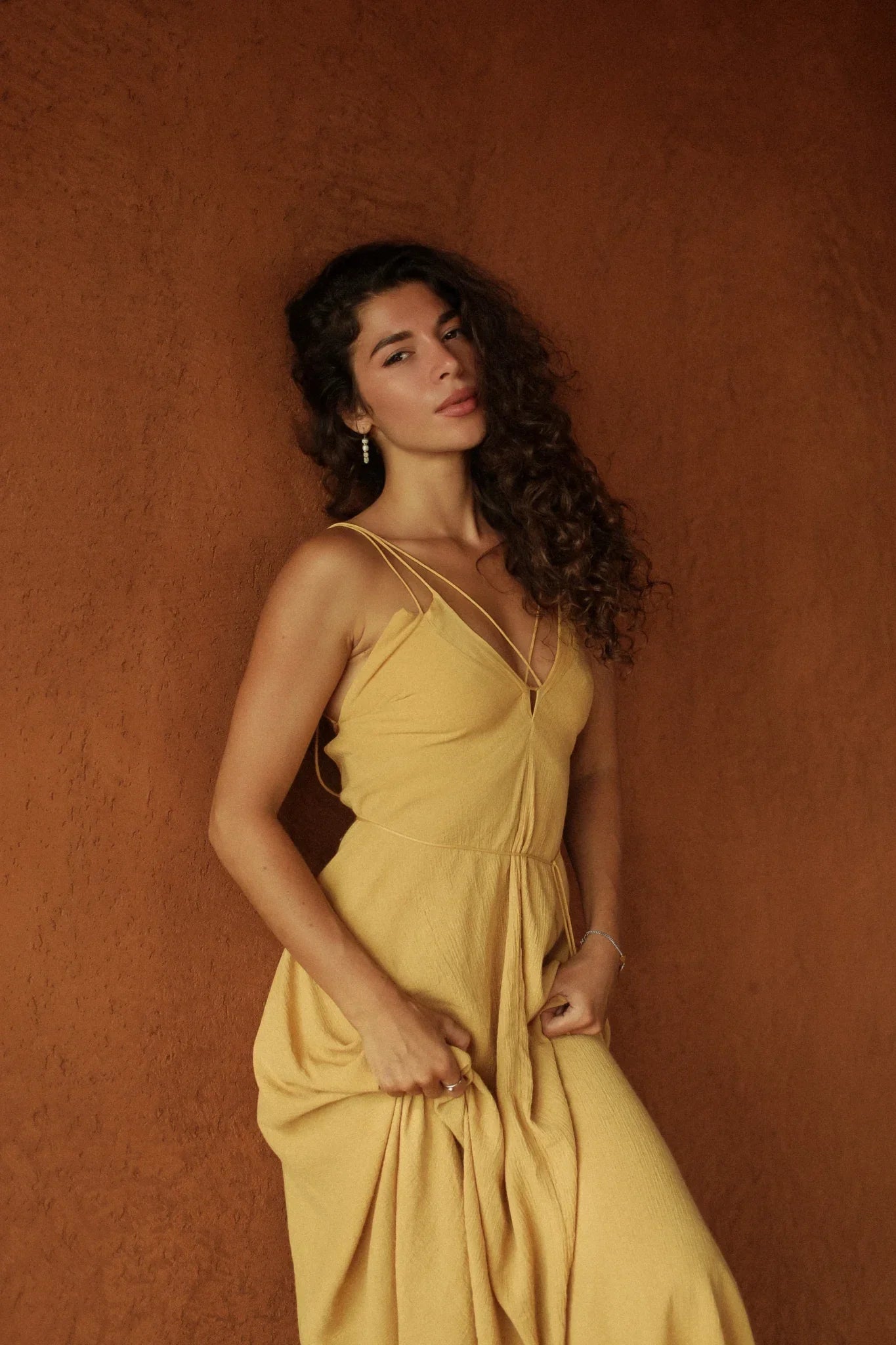 A woman with long curly hair wears the Powder Yellow Padma Dress by AYA Sacred Wear, a sleeveless boho backless dress, standing against a textured brown wall and gazing calmly at the camera while lightly holding her dress.
