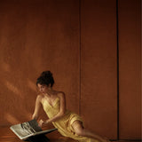 A woman in the Powder Yellow Padma Dress by AYA Sacred Wear sits on a patterned rug, leaning against wood, reading a large book in a warmly lit room with earthy brown walls.