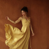 A woman in the Powder Yellow Padma Dress by AYA Sacred Wear stands barefoot against a textured brown wall, gently lifting the hem of her sleeveless boho backless dress and gazing downward with a serene expression.