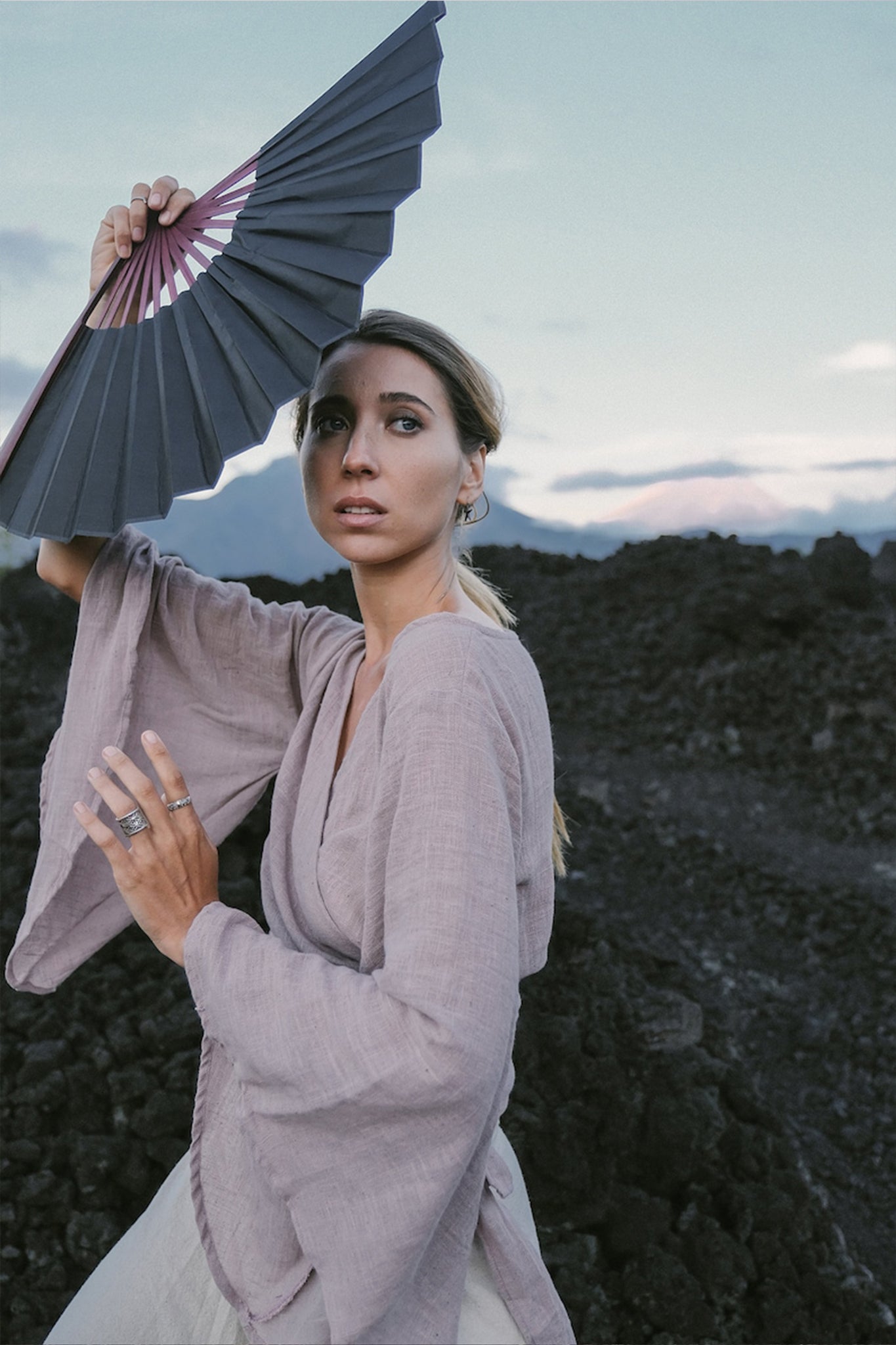 A woman wearing the Dry Lavender Kimono Cover-up Top Tunic from AYA Sacred Wear, crafted from raw organic cotton, holds a large hand fan above her head. She stands against a backdrop of dark volcanic rocks and a cloudy sky, embodying sustainable fashion amidst nature's dramatic canvas.