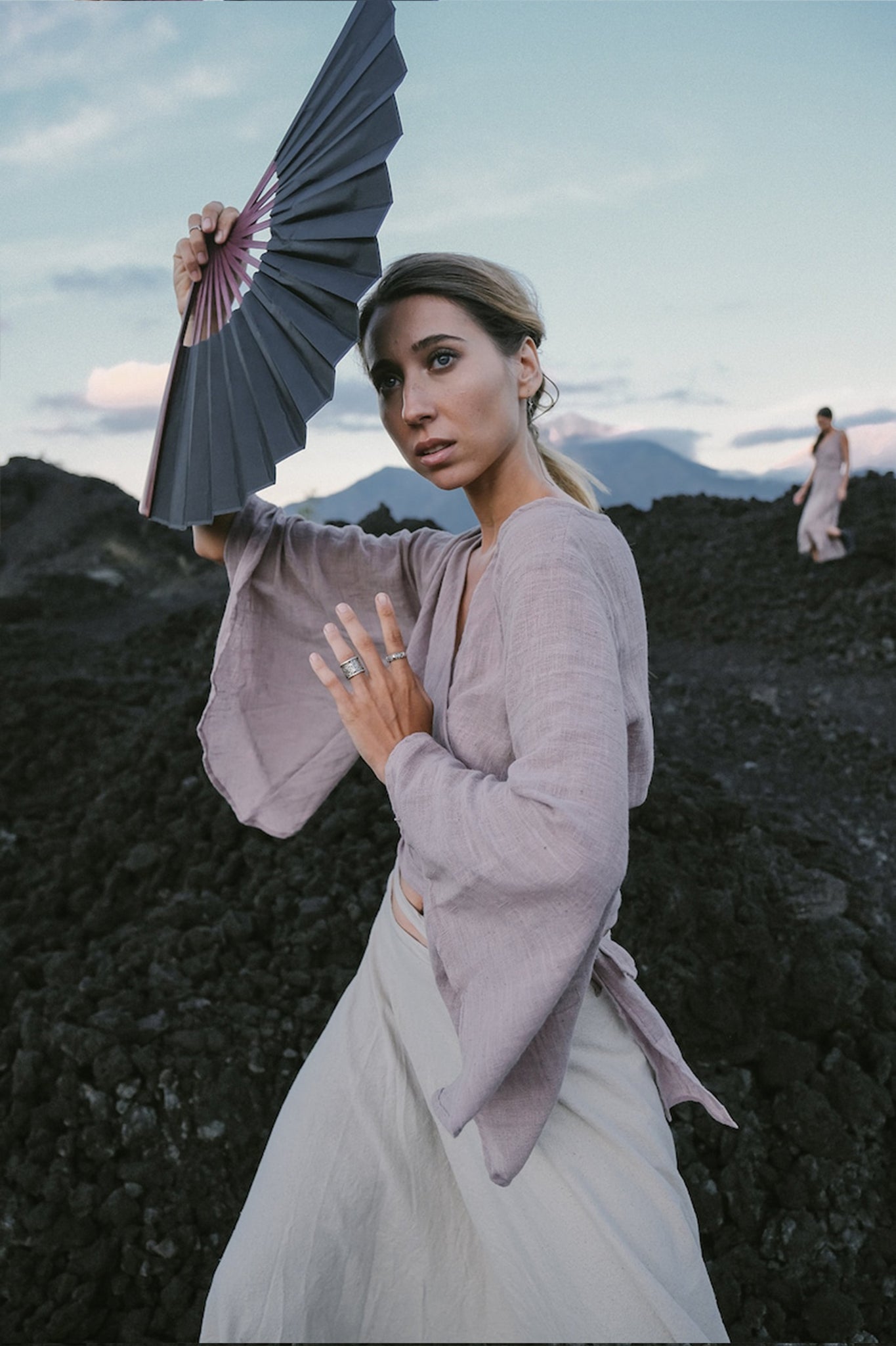 A woman wearing a light and breezy Dry Lavender Kimono Cover-up Top Tunic by AYA Sacred Wear holds a fan above her head as she stands on the dark, rocky terrain. The sky is partly cloudy, and another person can be seen in the background. The scene exudes a calm and natural vibe, showcasing sustainable fashion.