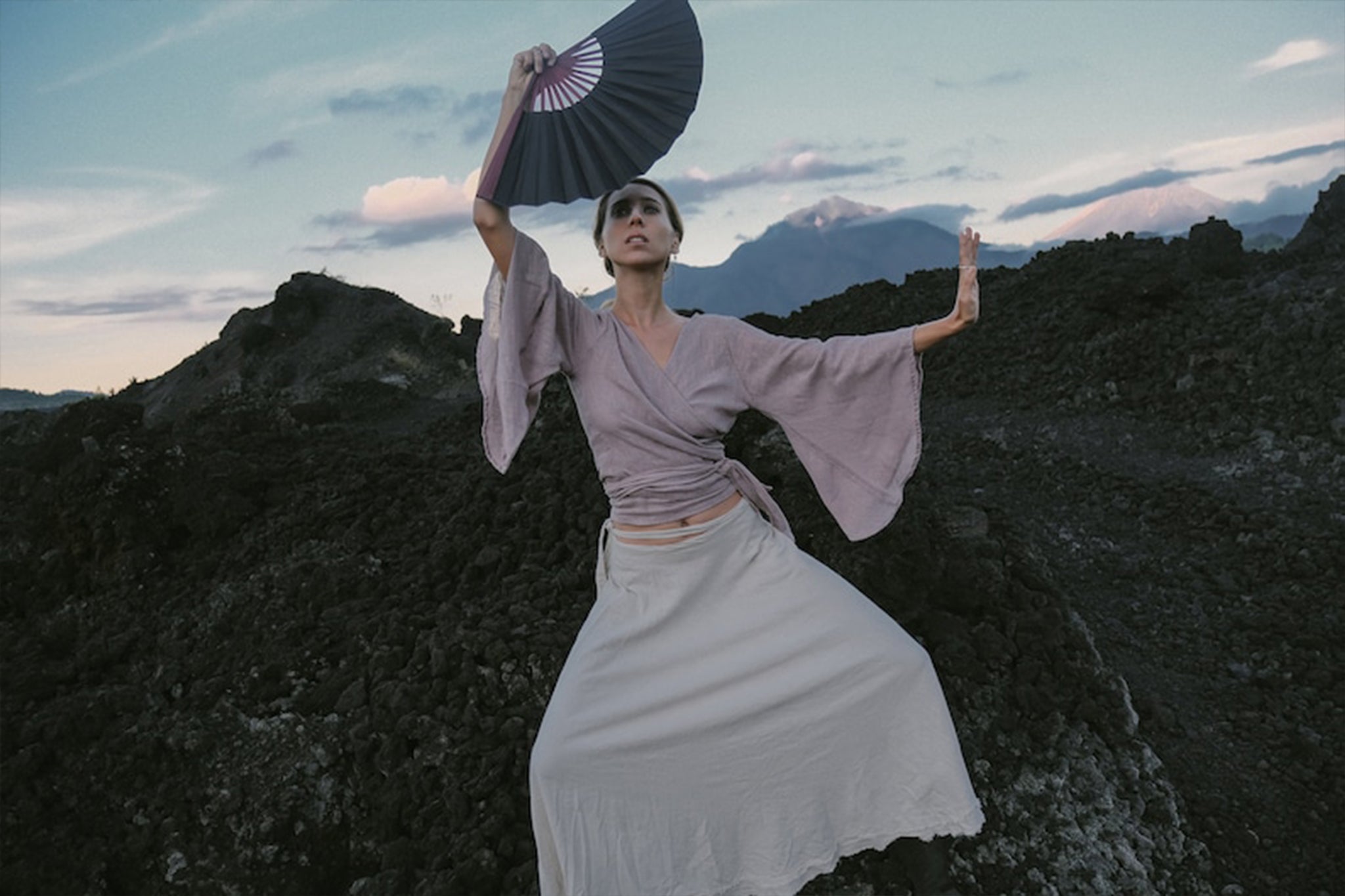 Standing dramatically on rocky terrain, an individual is seen wearing the Dry Lavender Kimono Cover-up Top Tunic from AYA Sacred Wear. Made of raw organic cotton, this flowing wrap blouse complements their pose, as they hold an open fan above their head. The cloudy sky and mountains in the background enhance the captivating appeal of sustainable fashion.