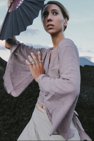 A person poses outdoors in a Dry Lavender Kimono Cover-up Top Tunic by AYA Sacred Wear, made from raw organic cotton. They hold a partially opened fan above their head, set against a backdrop of dark rocky terrain and rising mountains under a cloudy sky, capturing the essence of sustainable fashion.