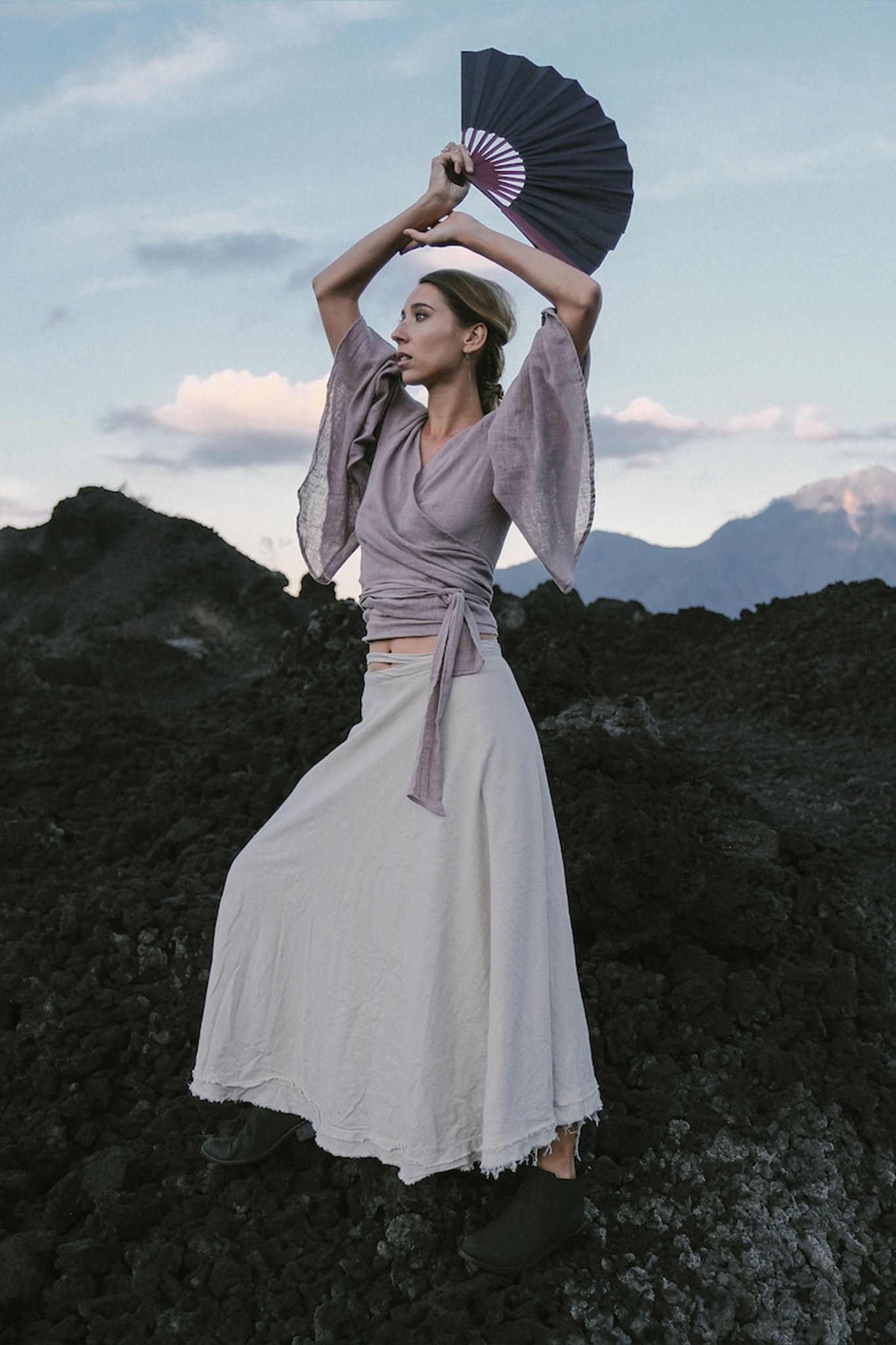 Amidst the dark rocky landscape, a person is elegantly dressed in a Dry Lavender Kimono Cover-up Top Tunic from AYA Sacred Wear. They hold a decorative fan aloft against the backdrop of distant mountains and cloudy skies, embodying the spirit of sustainable fashion.