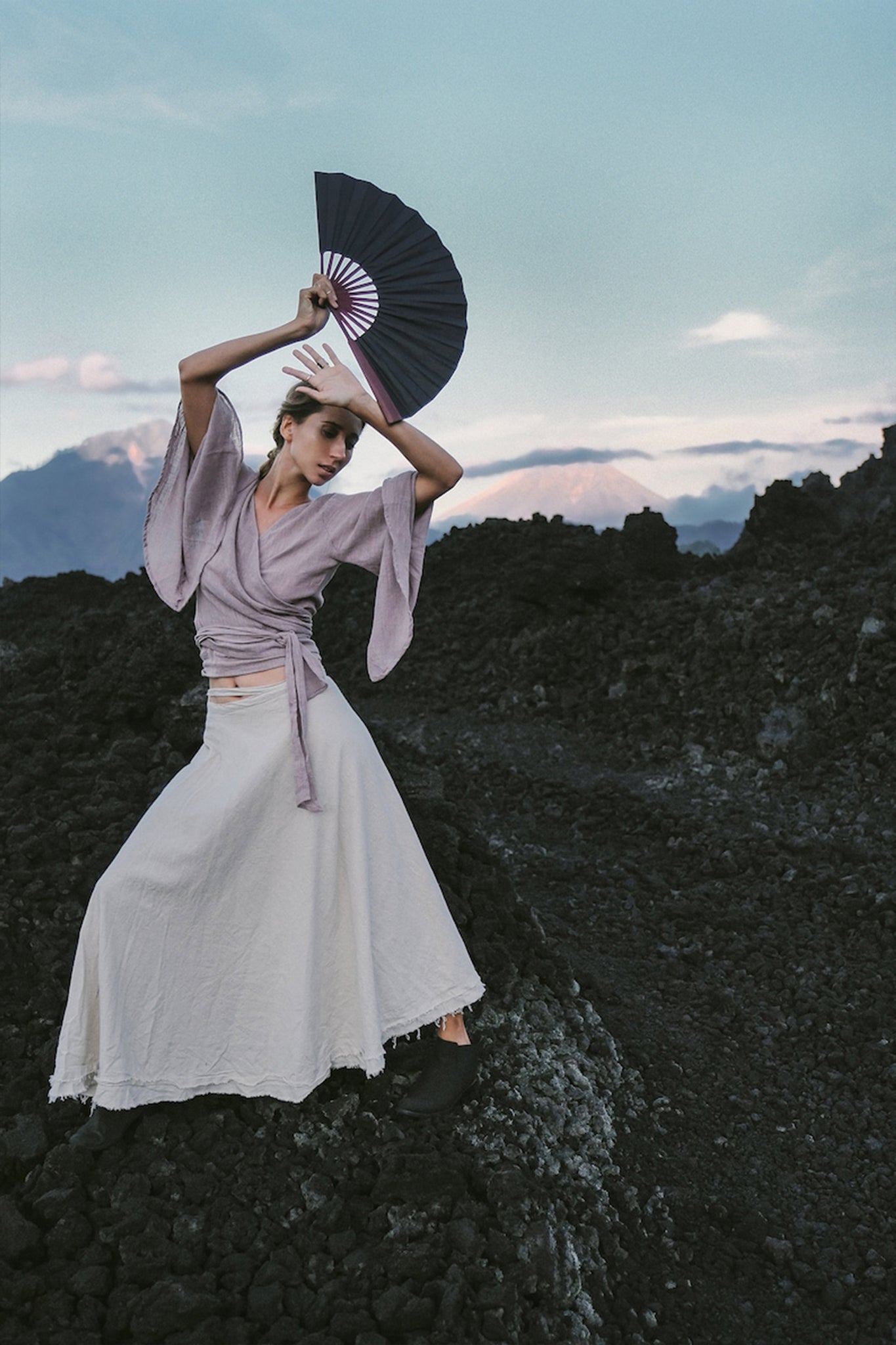 A person poses dramatically on rocky terrain, showcasing the Dry Lavender Kimono Cover-up Top Tunic from AYA Sacred Wear, holding a folded black fan. They wear a long, flowing white skirt. The background reveals a landscape with mountains under a blue sky, embodying the essence of sustainable fashion.