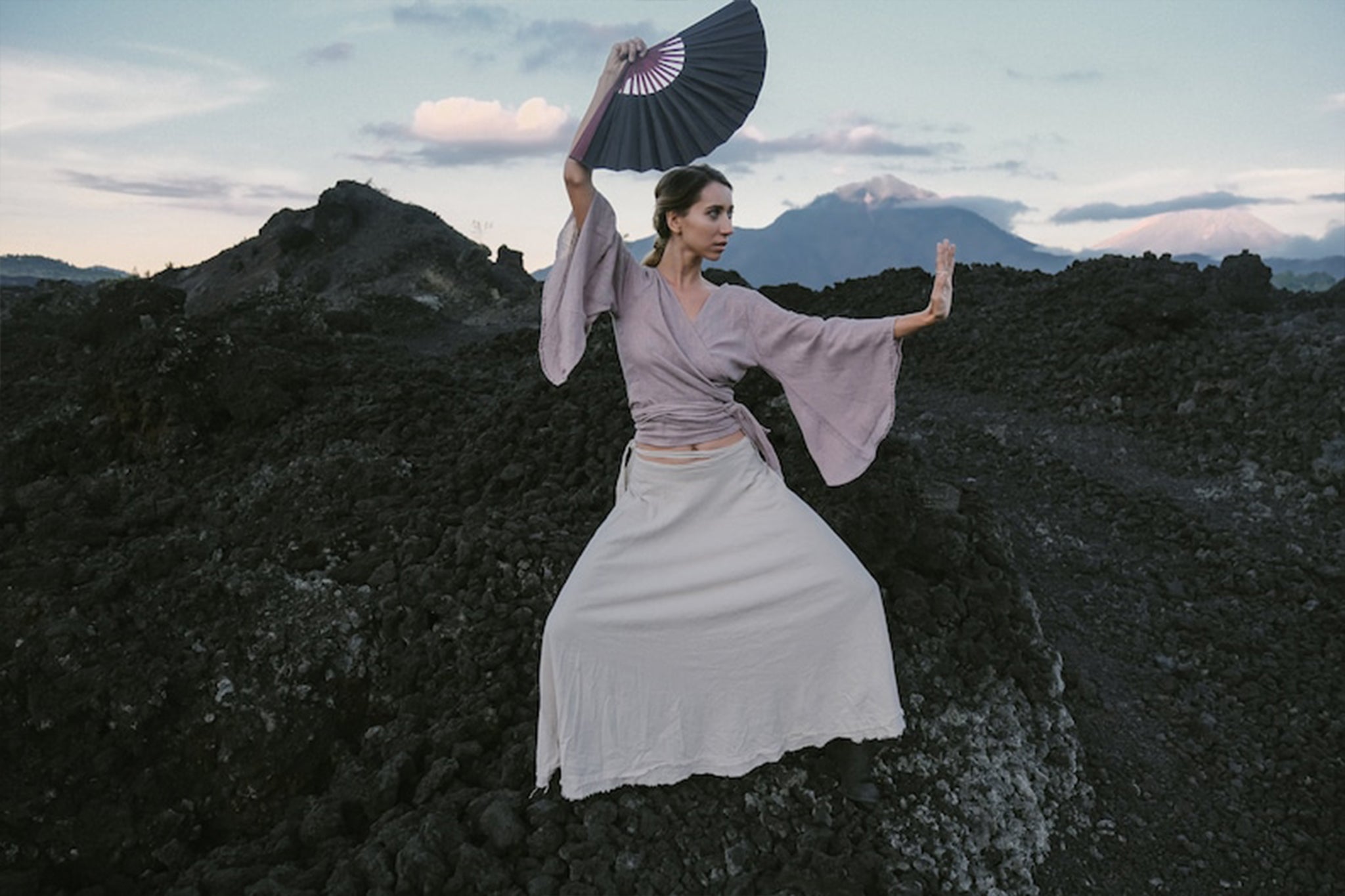 A person in traditional clothing poses dramatically on rocky terrain, holding an open fan. They are wearing the Dry Lavender Kimono Cover-up Top Tunic from AYA Sacred Wear, paired with a long beige skirt made from raw organic cotton. The background features dark rocks and distant mountains under a cloudy sky.