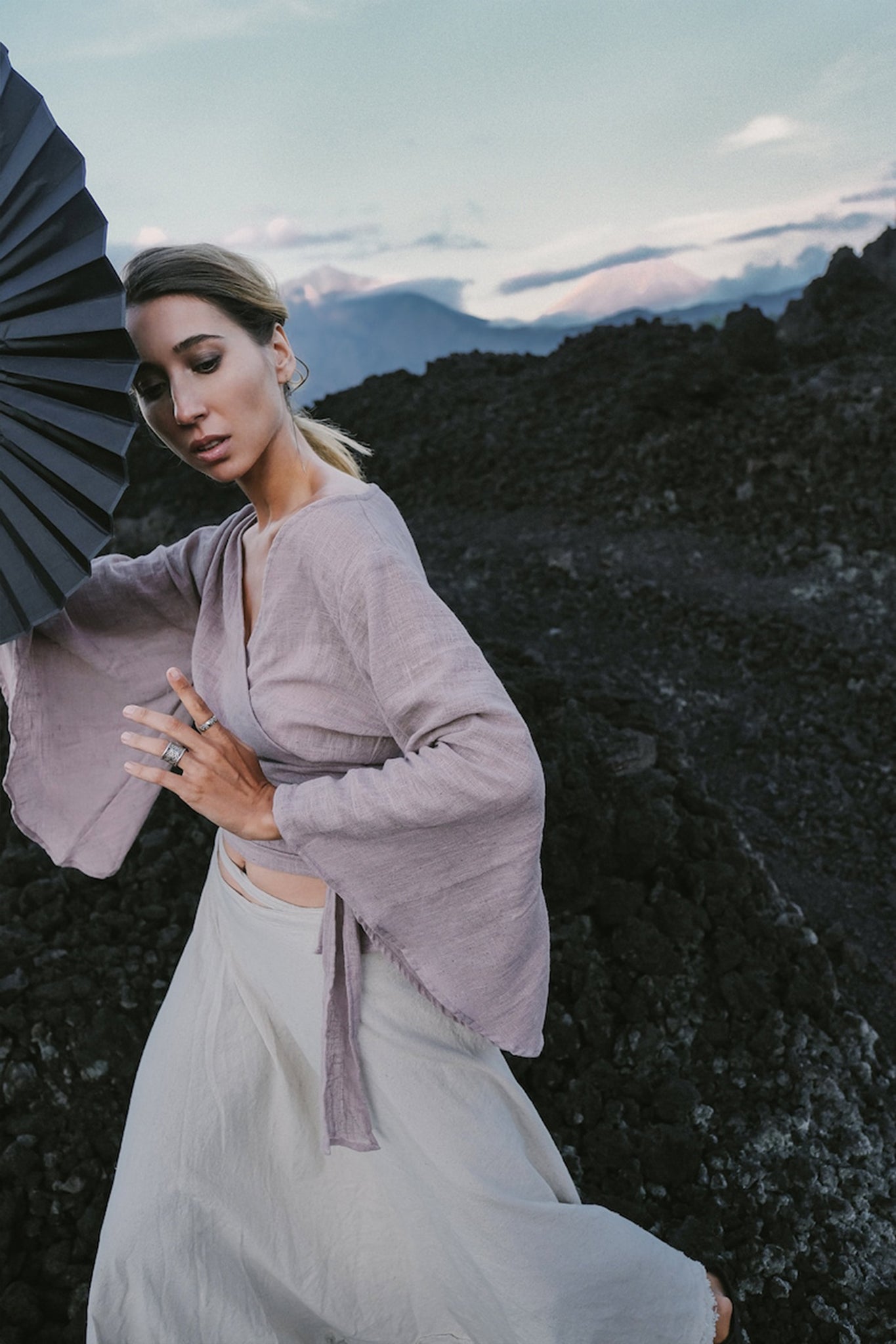 Wearing a Dry Lavender Kimono Cover-up Top Tunic by AYA Sacred Wear, a person poses gracefully with a black fan against a backdrop of dark, rocky terrain. The scene features partly cloudy skies and distant mountains, enhancing the dramatic yet serene atmosphere that reflects the essence of sustainable fashion.