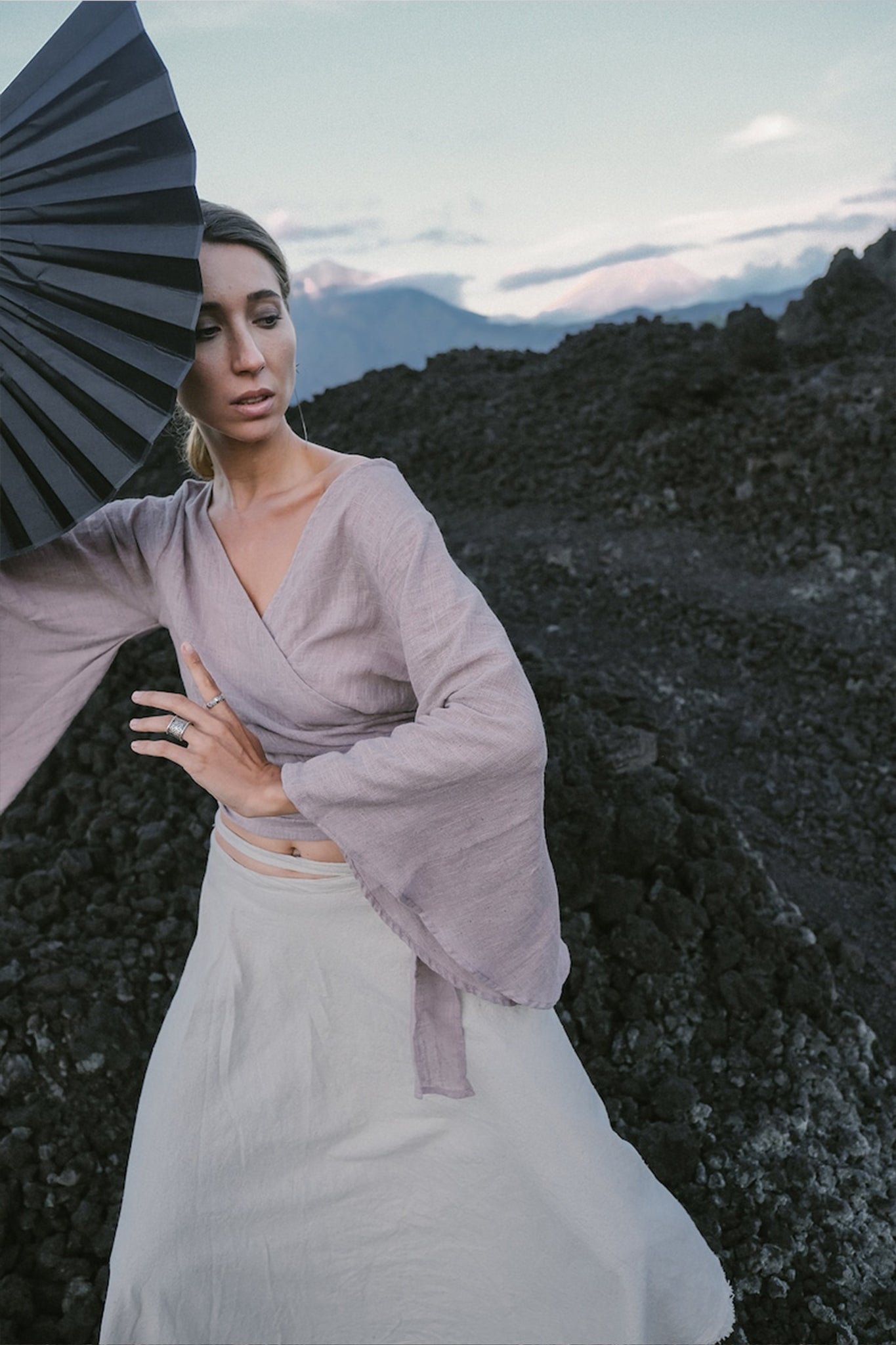 Dressed in an AYA Sacred Wear Dry Lavender Kimono Cover-up Top Tunic and a beige skirt, she embodies sustainable fashion while holding a black fan on a rocky landscape, mountains framing her figure. Her gaze is directed downward as the sky unfolds its soft, pastel hues behind her.