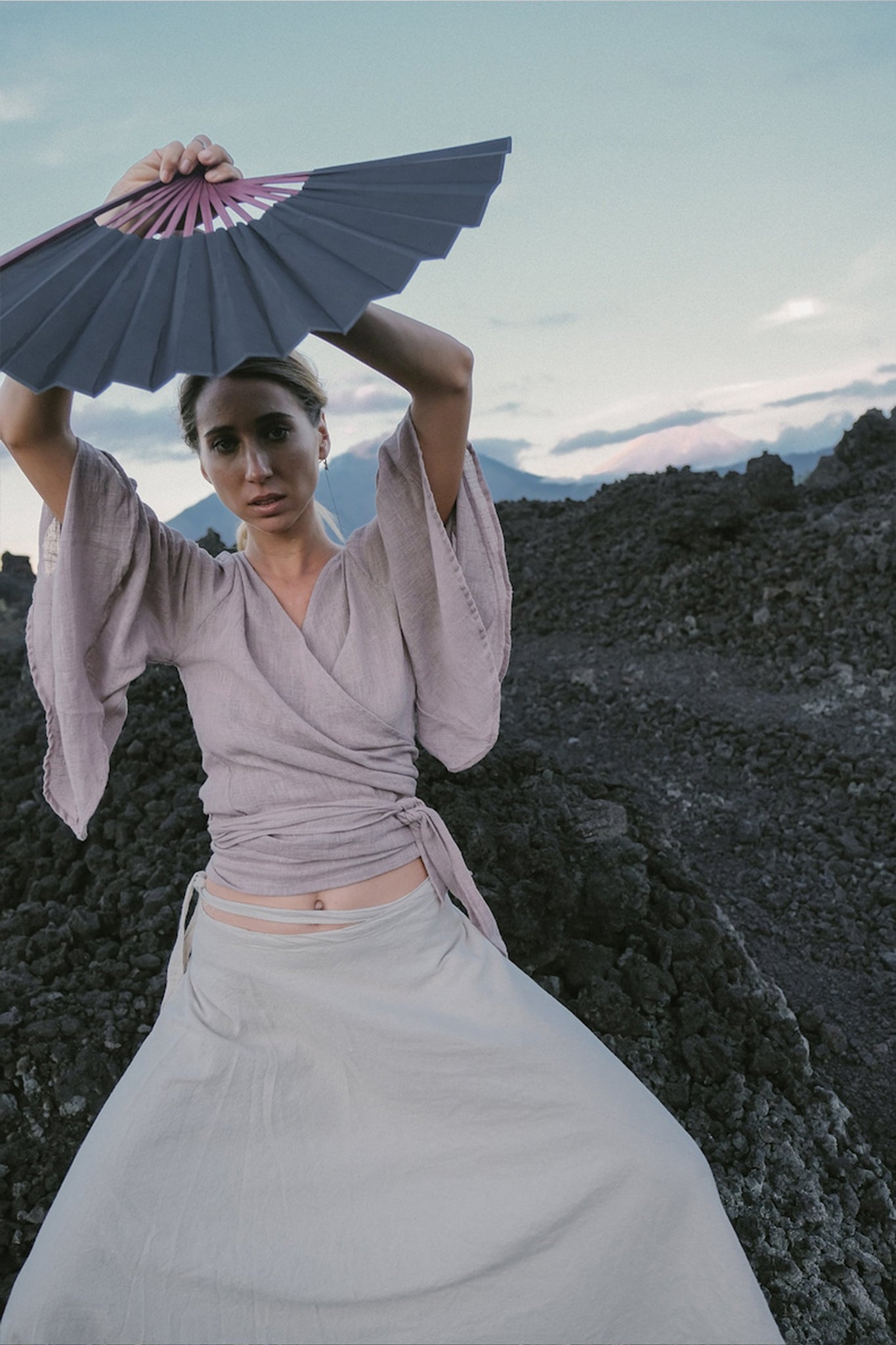 Dressed in a Dry Lavender Kimono Cover-up Top Tunic from AYA Sacred Wear, a woman sits on a rocky landscape wearing a long beige skirt made from raw organic cotton. She holds a black and pink fan above her head against the backdrop of clear skies with soft clouds and distant mountains, capturing the essence of sustainable fashion.