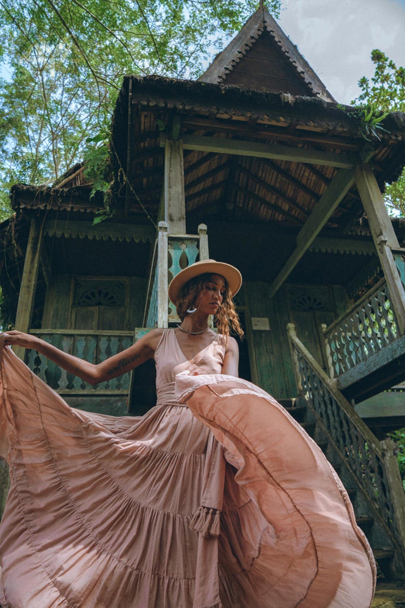 A woman in an elegant Dusty Pink Bohemian Dress from AYA Sacred Wear, complete with a hat, poses gracefully before a traditional rustic wooden house amidst lush greenery. The house is adorned with intricate woodwork and boasts a steep, pointed roof.