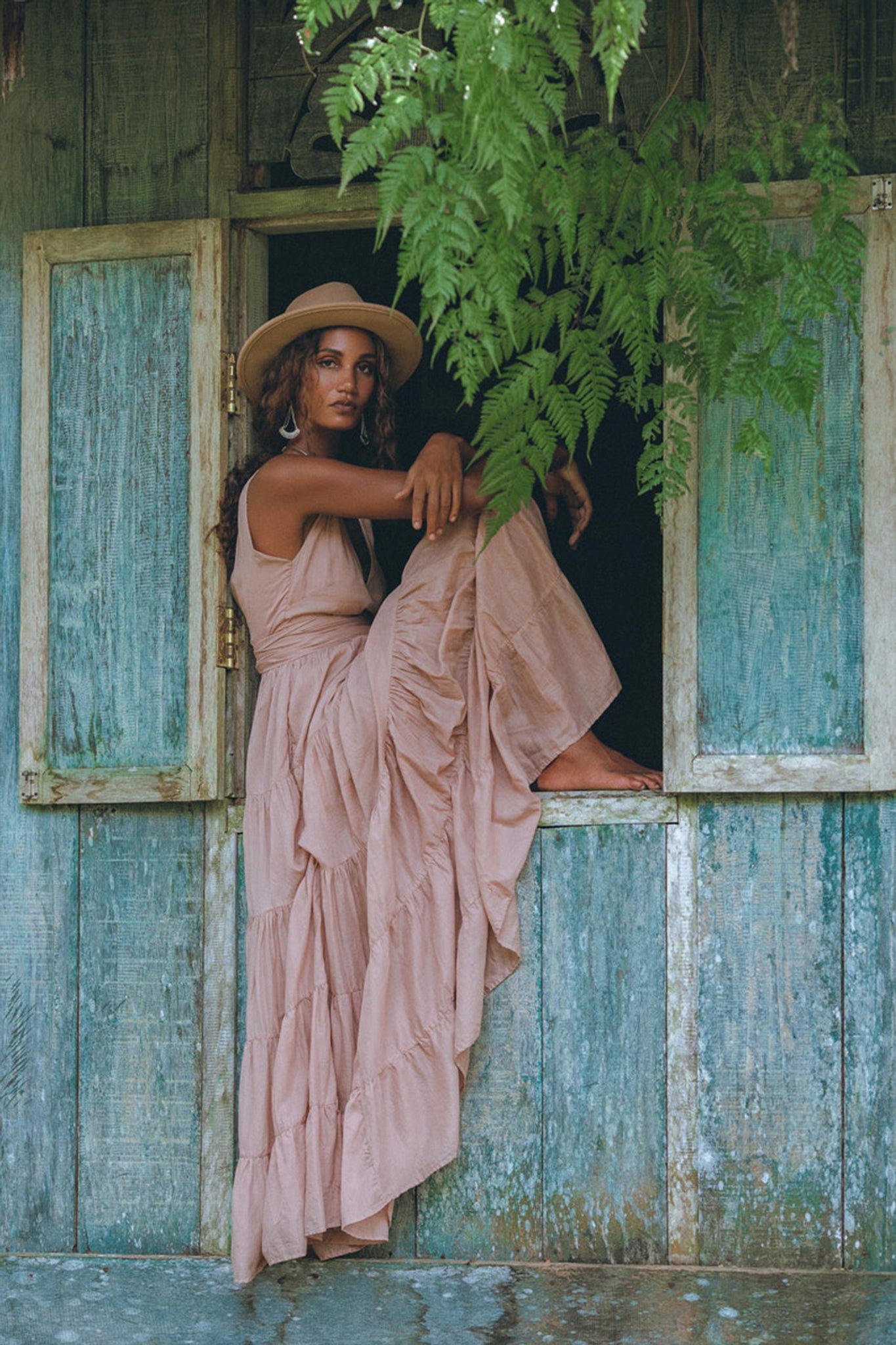 A woman in an elegant Dusty Pink Bohemian Dress by AYA Sacred Wear sits within a rustic wooden window frame. Greenery hangs above her, and the weathered window shutters add to the organic, vintage charm of the scene.