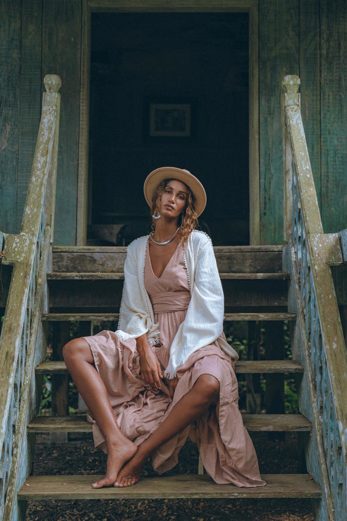 A person sits on wooden steps in a relaxed pose, wearing a wide-brimmed hat, an AYA Sacred Wear Dusty Pink Bohemian Dress and a light cardigan. The setting appears rustic with weathered wooden details and a dark doorway in the background.