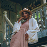 A woman wearing the Dusty Pink Bohemian Dress from AYA Sacred Wear, combined with a wide-brimmed hat, poses gracefully on wooden steps. She stands before a rustic building adorned with intricate carvings, enveloped by lush greenery. The atmosphere exudes serenity and style.