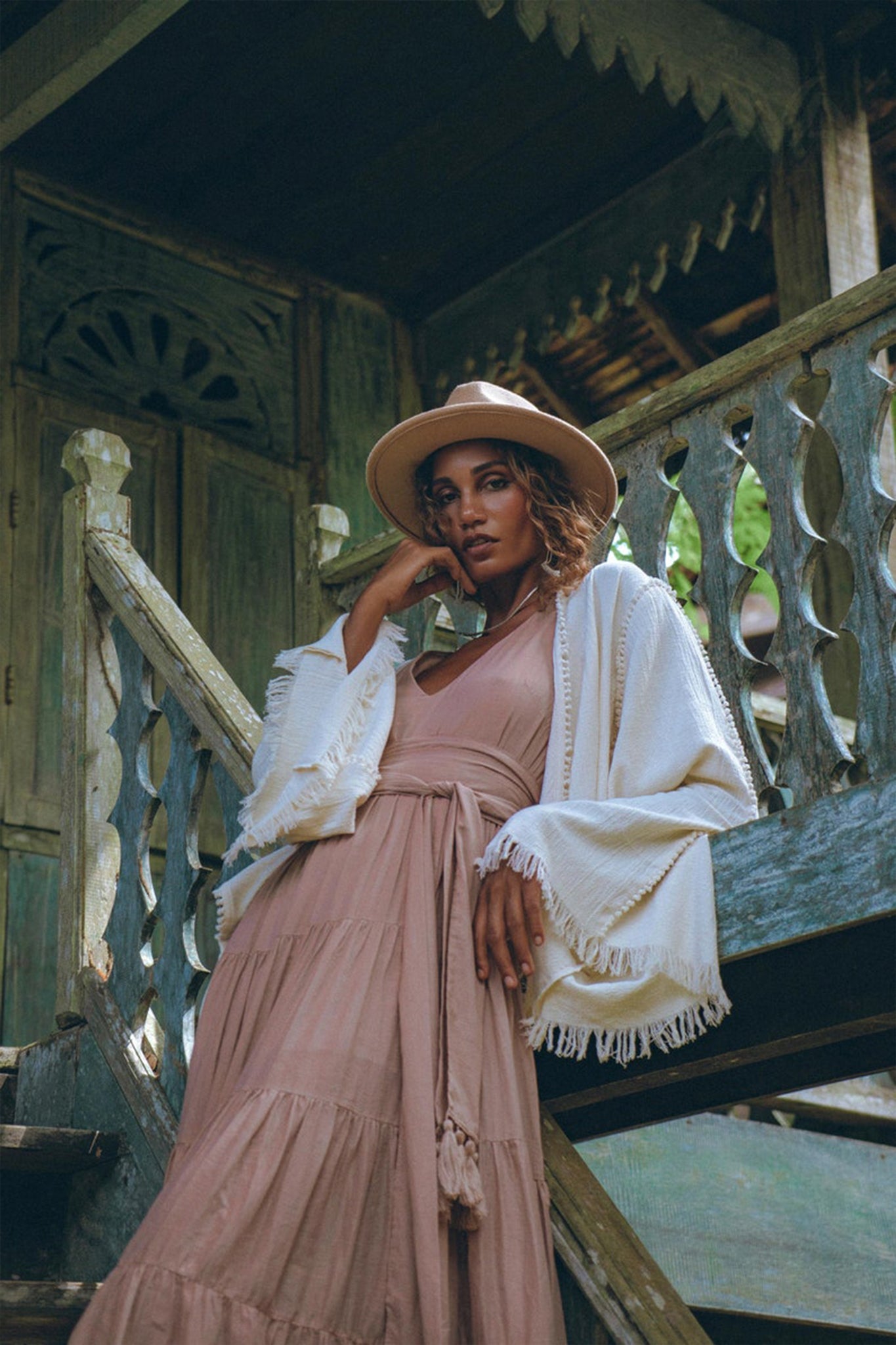 A woman wearing the Dusty Pink Bohemian Dress from AYA Sacred Wear, combined with a wide-brimmed hat, poses gracefully on wooden steps. She stands before a rustic building adorned with intricate carvings, enveloped by lush greenery. The atmosphere exudes serenity and style.