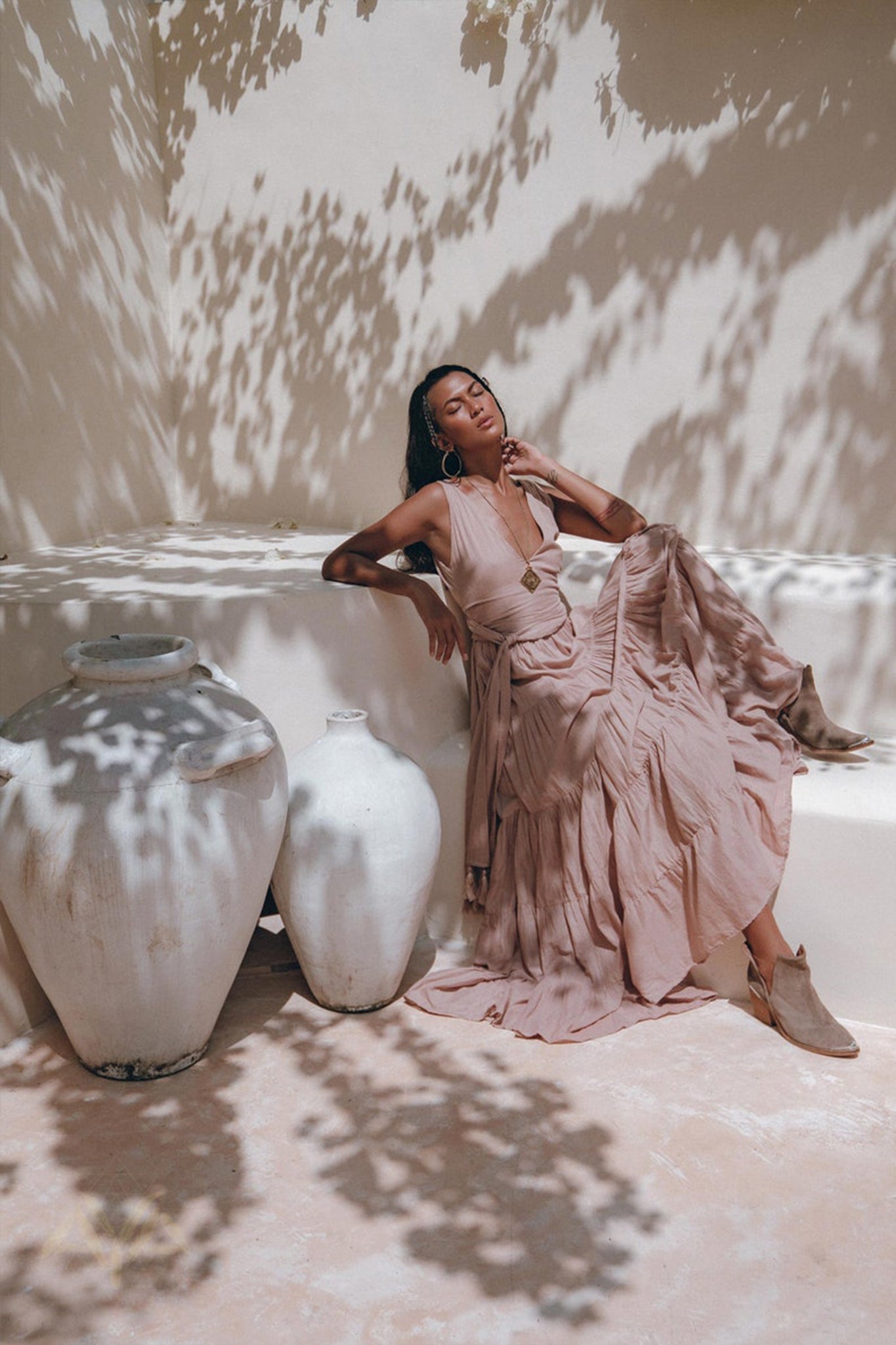 A woman wearing a Dusty Pink Bohemian Dress from AYA Sacred Wear, paired with boots, sits on a white bench and leans against a wall. Large white pottery jars are nearby, while dappled sunlight and leaf shadows create patterns across the scene.