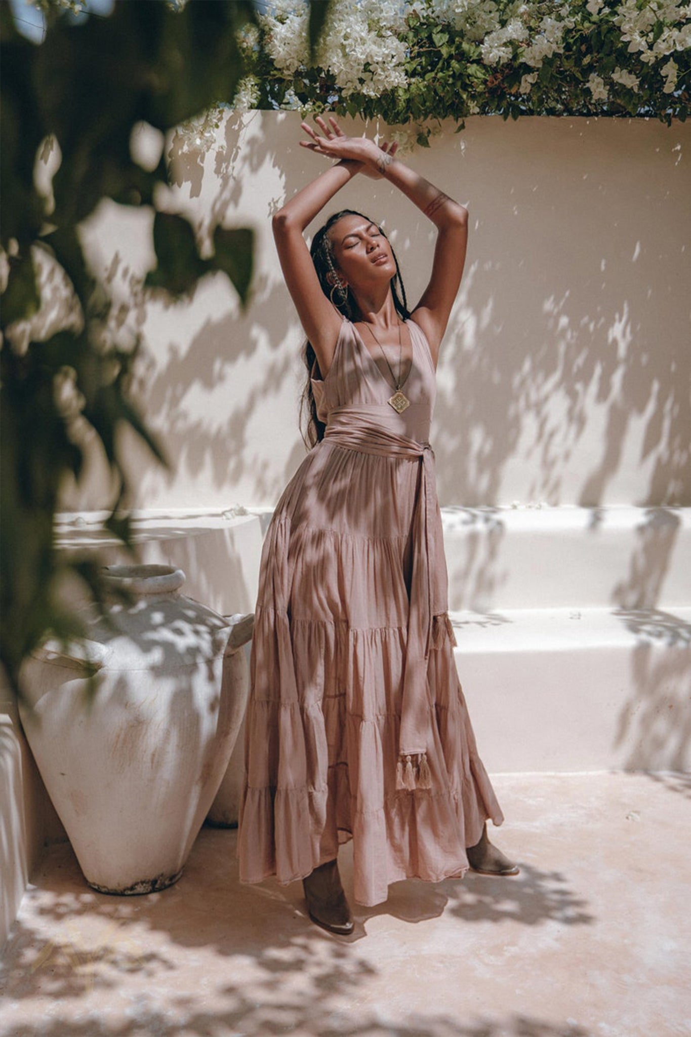 In a sunlit garden, a woman strikes an elegant pose in an AYA Sacred Wear Dusty Pink Bohemian Dress, featuring its flowing silhouette and A-line belted design. With arms raised gracefully, she is surrounded by large white vases and the dappled light and shadows cast by leaves and flowers overhead.