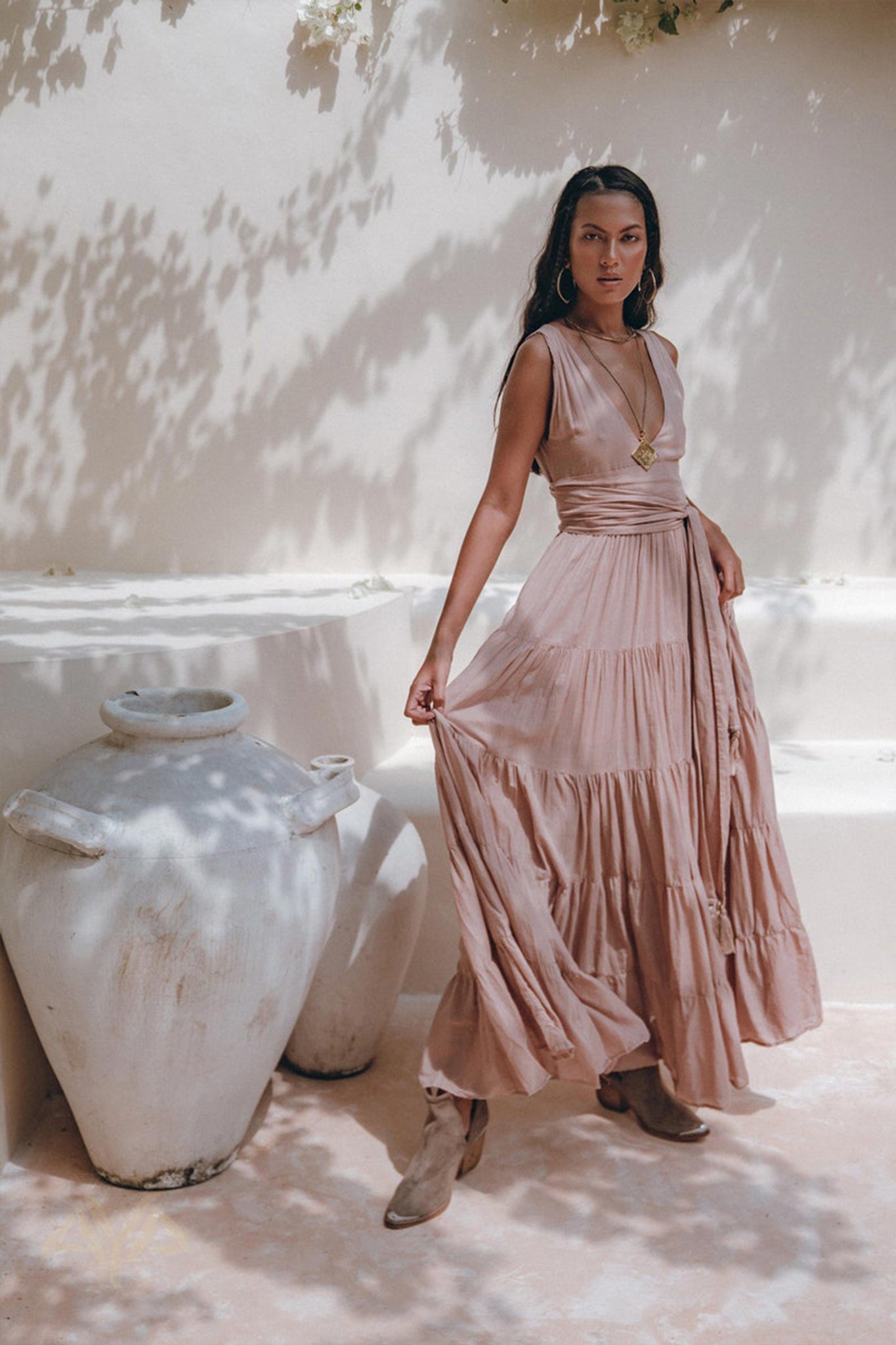 A woman elegantly poses outdoors in a flowing, sleeveless Dusty Pink Bohemian Dress by AYA Sacred Wear, surrounded by rustic pottery. The sunny scene casts soft shadows from nearby foliage. Complementing her look with large hoop earrings, a necklace, and ankle boots, she embodies a Boho Gypsy vibe.