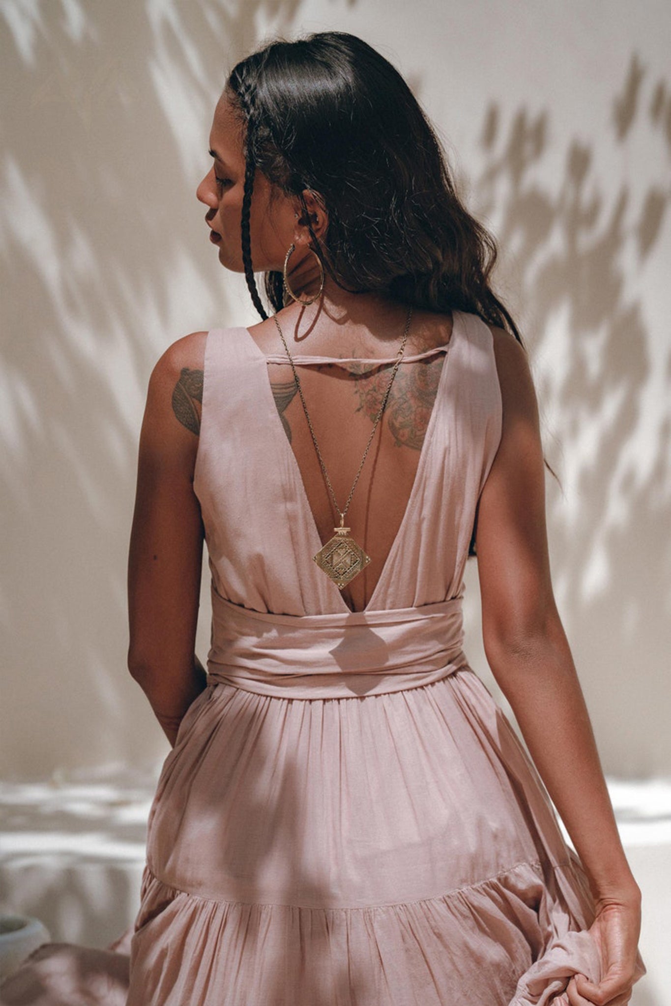 A woman with long hair stands with her back to the camera, showcasing a Dusty Pink Bohemian Dress from AYA Sacred Wear. The dress features a V-shaped back that reveals a tattoo on her shoulder. She accessorizes with a long necklace adorned with a large pendant, while dappled sunlight plays patterns around her.