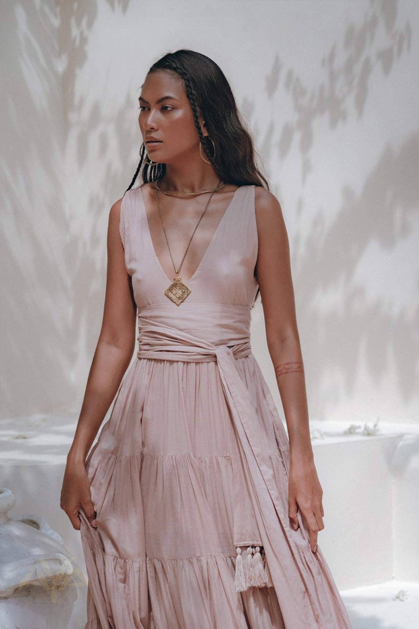 A woman in a long, sleeveless, dusty pink Bohemian dress from AYA Sacred Wear stands outdoors. Her dark hair flows beside large hoop earrings and a pendant necklace. The organic cotton fabric catches dappled sunlight amid the soft shadows of leaves.