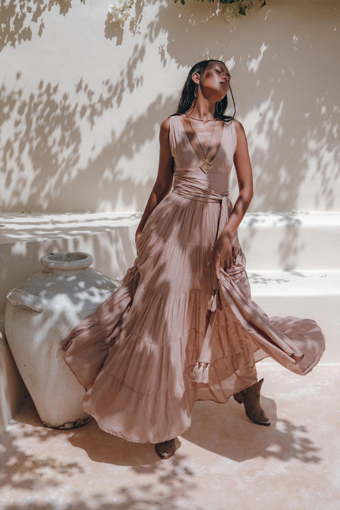 A woman stands gracefully outdoors against a sunlit wall, posing in the Dusty Pink Bohemian Dress by AYA Sacred Wear. The flowing A-line belted dress creates delicate dappled shadows, complemented by her large hoop earrings and pendant necklace. Nearby, a large white vase adds to the serene ambiance of the scene.