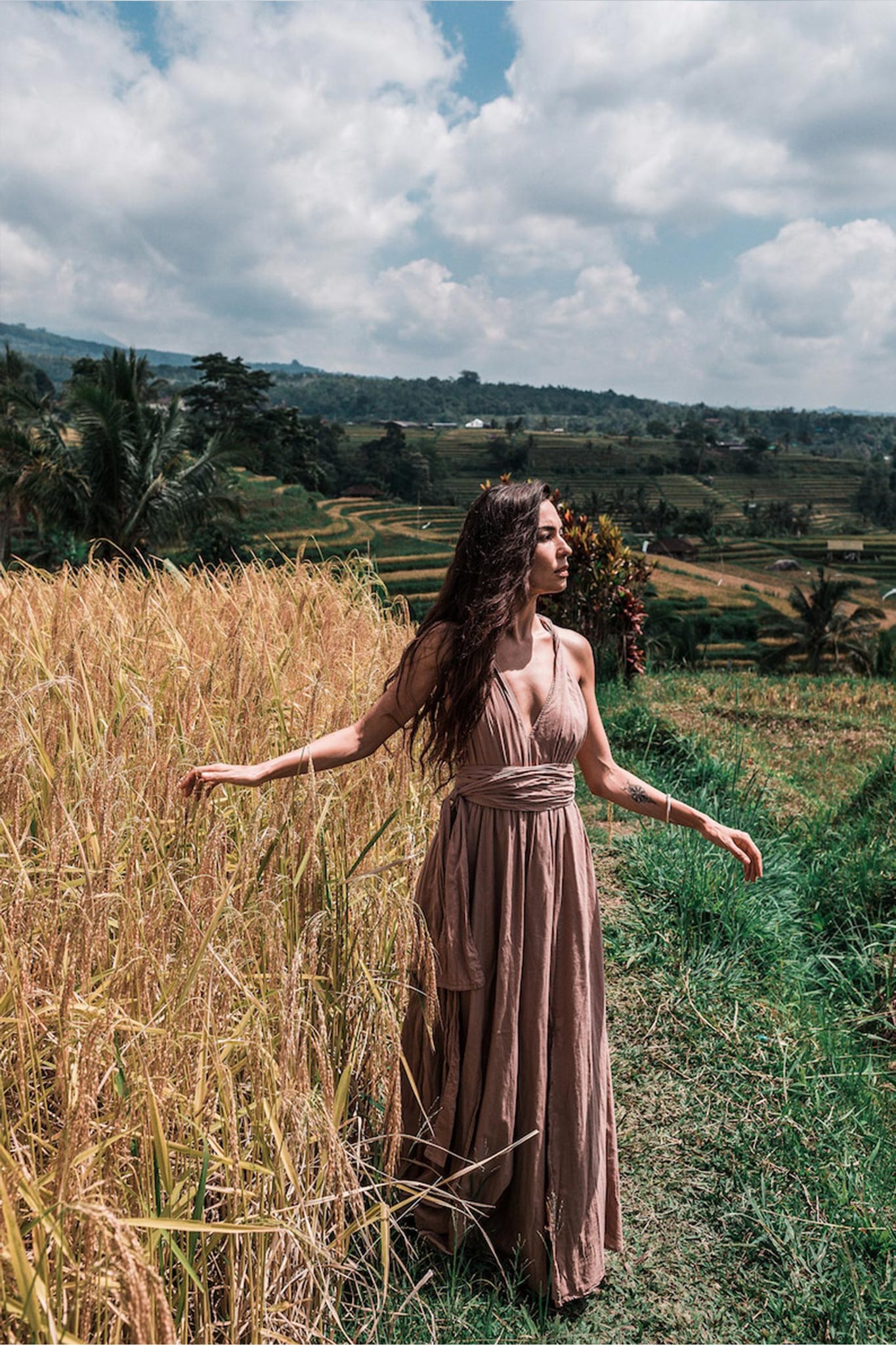 A woman wearing the Dusty Pink Boho Bridesmaid Dress Convertible by AYA Sacred Wear glides through a lush, green and golden field under a partly cloudy sky. Trees and hills provide a charming backdrop, completing the rural, scenic landscape.