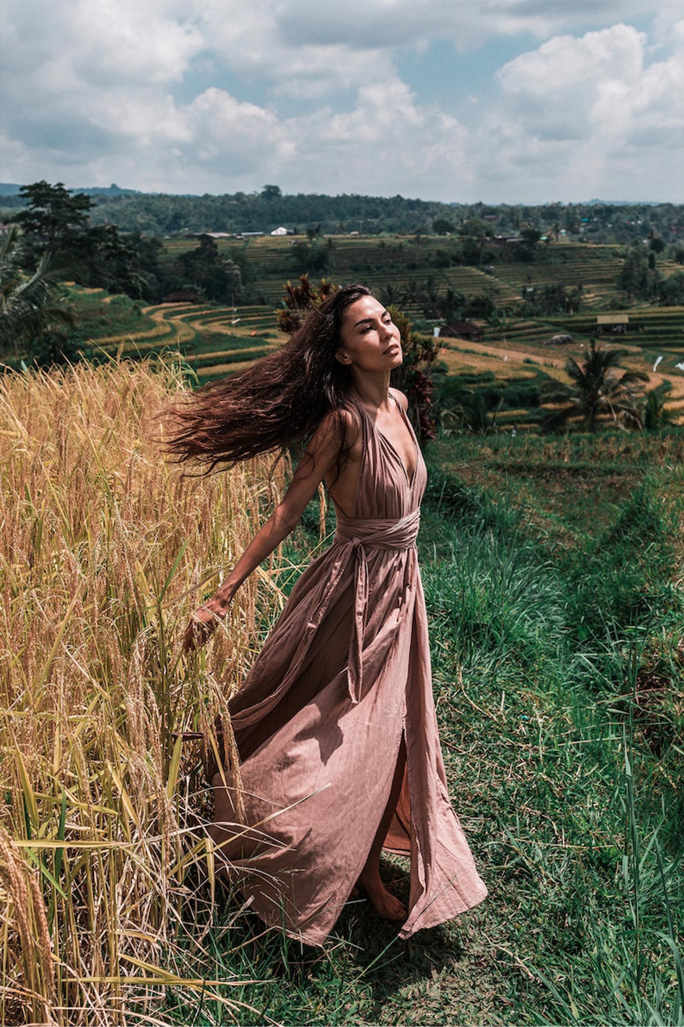 A woman in a Dusty Pink Boho Bridesmaid Dress Convertible from AYA Sacred Wear stands in a lush green field with terraces, basking in the sunlight. Her long hair blows in the wind as she gracefully poses, surrounded by nature under a partly cloudy sky.