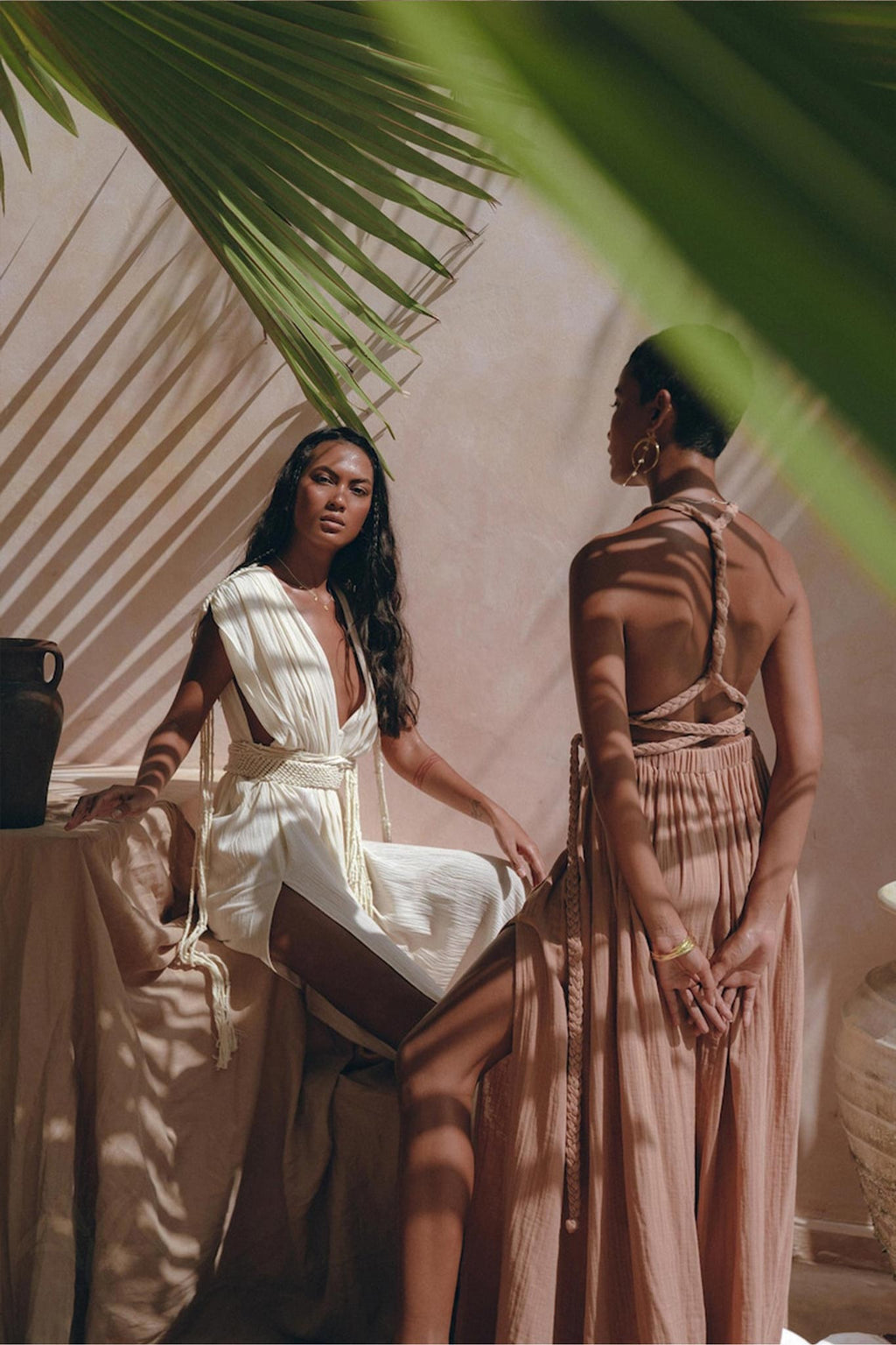 Two women in elegant dresses pose amid natural elements. One sits gracefully in a beautiful boho bridesmaid dress; the other stands, evoking a Greek goddess, wearing AYA Sacred Wear's Dusty Pink Greek Goddess Dress made from organic cotton, with her back to the camera. Palm shadows and sunlit surroundings create a serene ambiance.