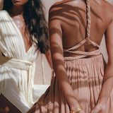 Two women are adorned in stylish, light-colored dresses. The woman in the foreground is wearing the Dusty Pink Greek Goddess Dress from AYA Sacred Wear, characterized by its braided straps and Grecian style. Meanwhile, the woman behind her wears a white wrap bridesmaid dress. Sunlight filters through to create leaf-shaped shadows across their dresses.