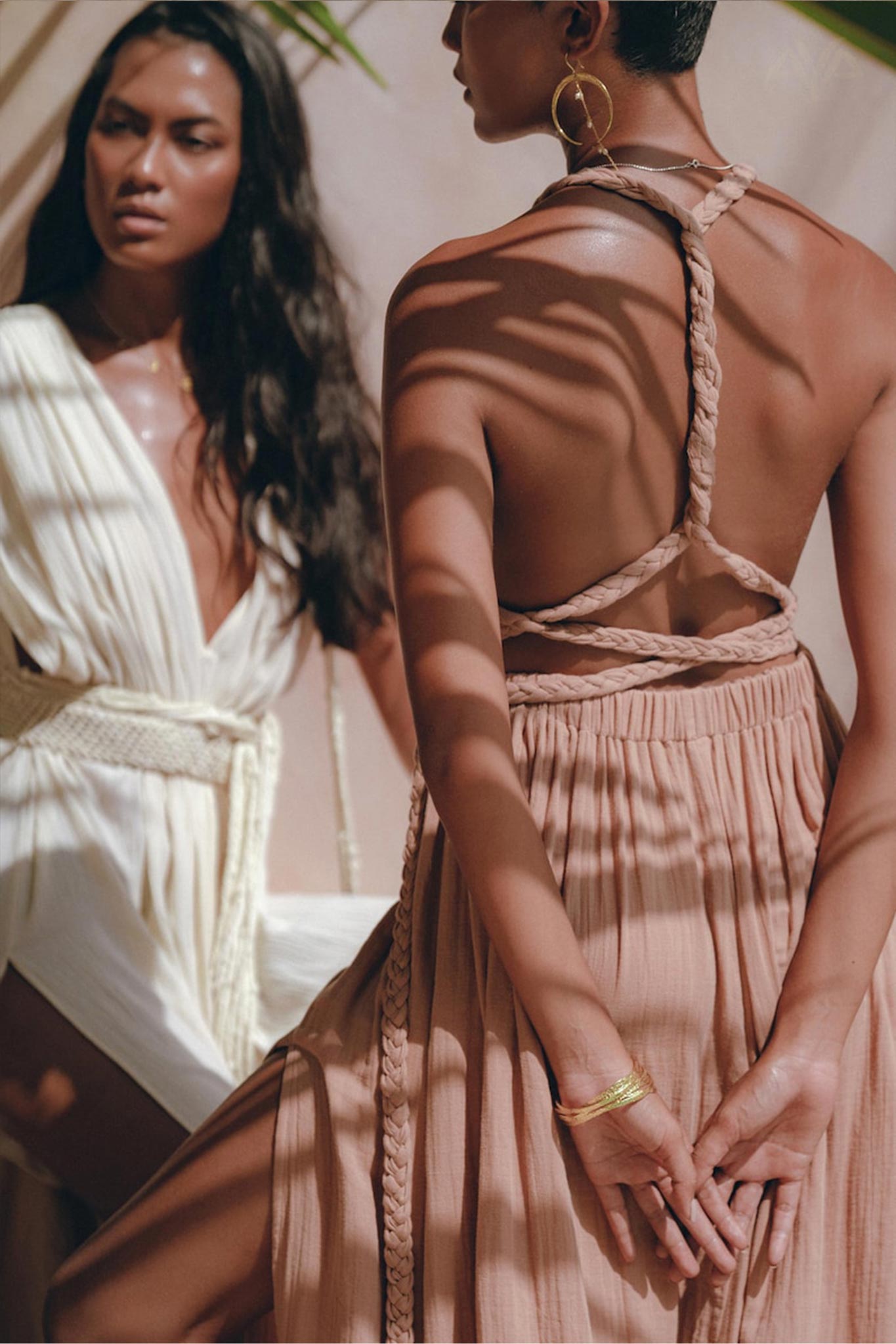 Two women are adorned in stylish, light-colored dresses. The woman in the foreground is wearing the Dusty Pink Greek Goddess Dress from AYA Sacred Wear, characterized by its braided straps and Grecian style. Meanwhile, the woman behind her wears a white wrap bridesmaid dress. Sunlight filters through to create leaf-shaped shadows across their dresses.