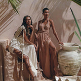 Two women pose in an earthy, desert-inspired setting. One is wearing a flowing boho bridesmaid dress paired with boots while sitting on a draped surface. The other stands beside a large ceramic vase, showcasing the Dusty Pink Greek Goddess Dress from AYA Sacred Wear. Large green leaves frame the scene beautifully.