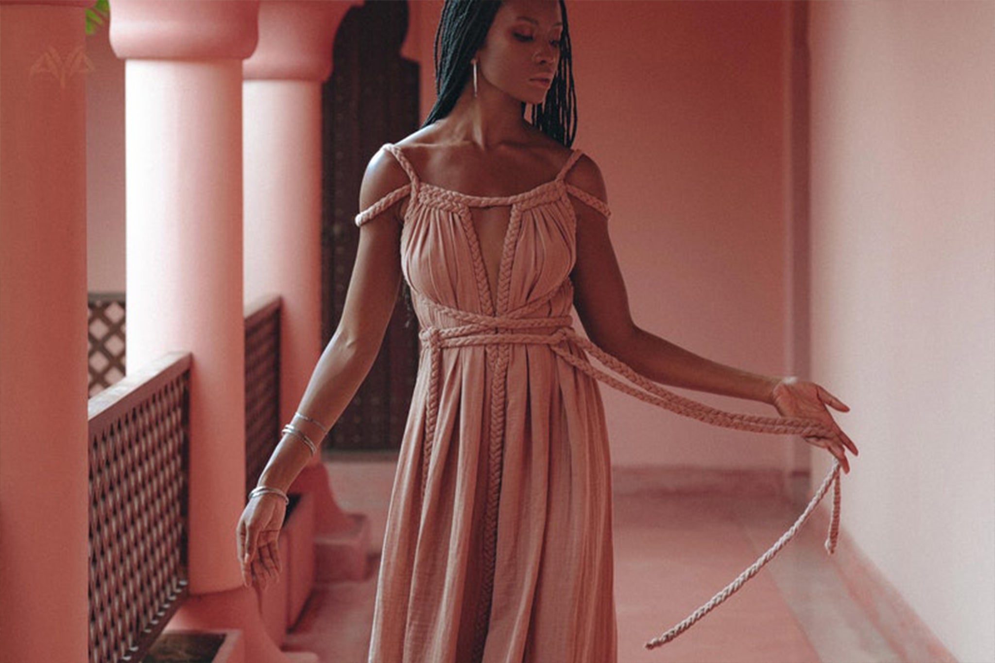 A person with long braided hair stands in a hallway with pink walls and columns, wearing the Dusty Pink Greek Goddess Prom Boho Dress by AYA Sacred Wear. They look down thoughtfully, holding one of the dress ties in their hand, capturing an elegant and contemplative posture that truly embodies the essence of this boho-inspired attire.