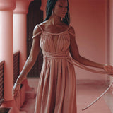 A person with long braided hair stands in a hallway with pink walls and columns, wearing the Dusty Pink Greek Goddess Prom Boho Dress by AYA Sacred Wear. They look down thoughtfully, holding one of the dress ties in their hand, capturing an elegant and contemplative posture that truly embodies the essence of this boho-inspired attire.