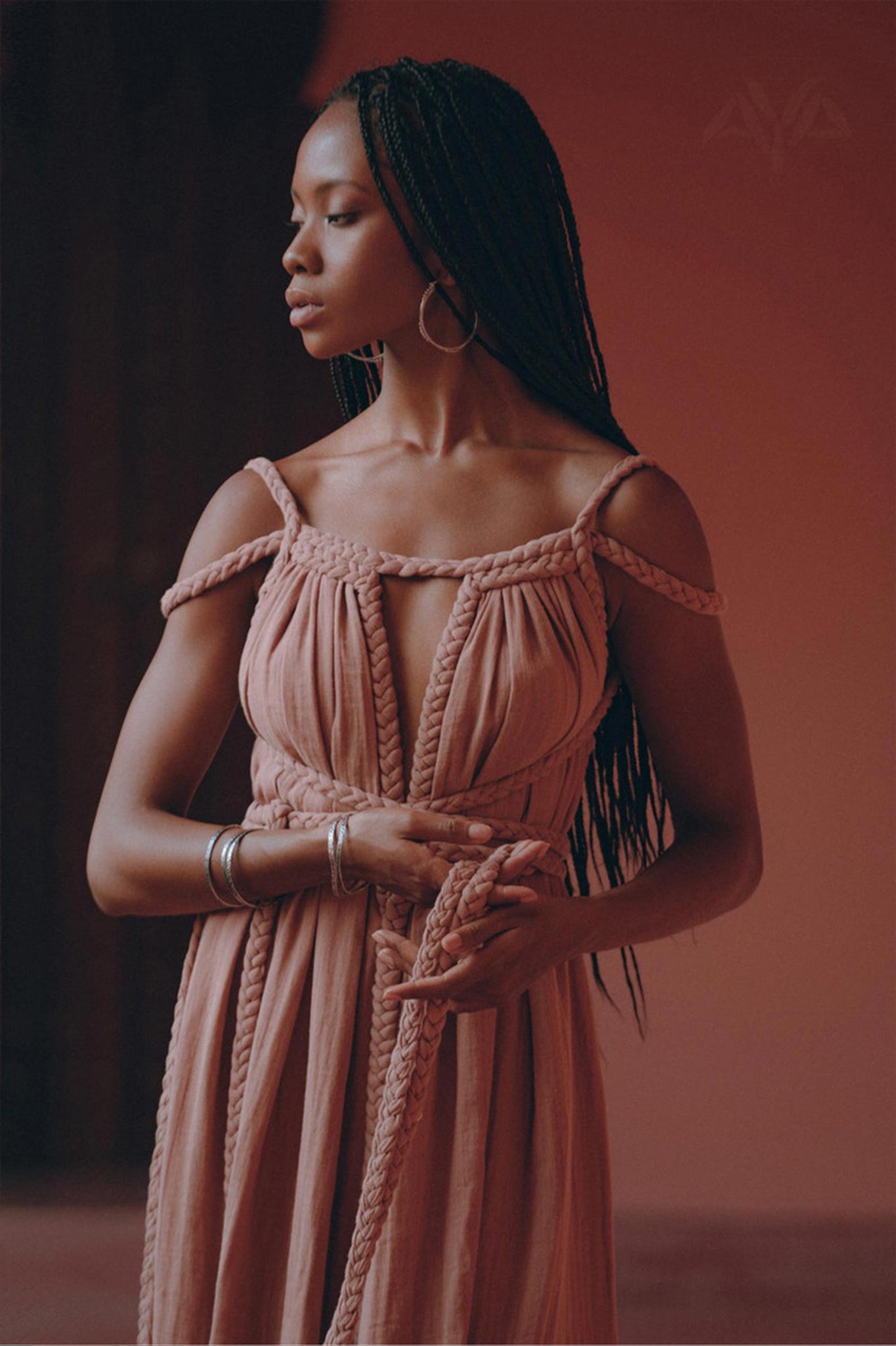 A woman with long braided hair stands gracefully, dressed in a flowy off-shoulder Dusty Pink Greek Goddess Prom Boho Dress by AYA Sacred Wear, complemented by hoop earrings, against a warm, peach-colored background. She appears contemplative as she holds the fabric of her dress in her hands.