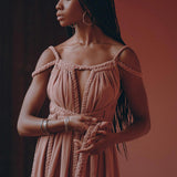 A woman with long braided hair stands gracefully, dressed in a flowy off-shoulder Dusty Pink Greek Goddess Prom Boho Dress by AYA Sacred Wear, complemented by hoop earrings, against a warm, peach-colored background. She appears contemplative as she holds the fabric of her dress in her hands.