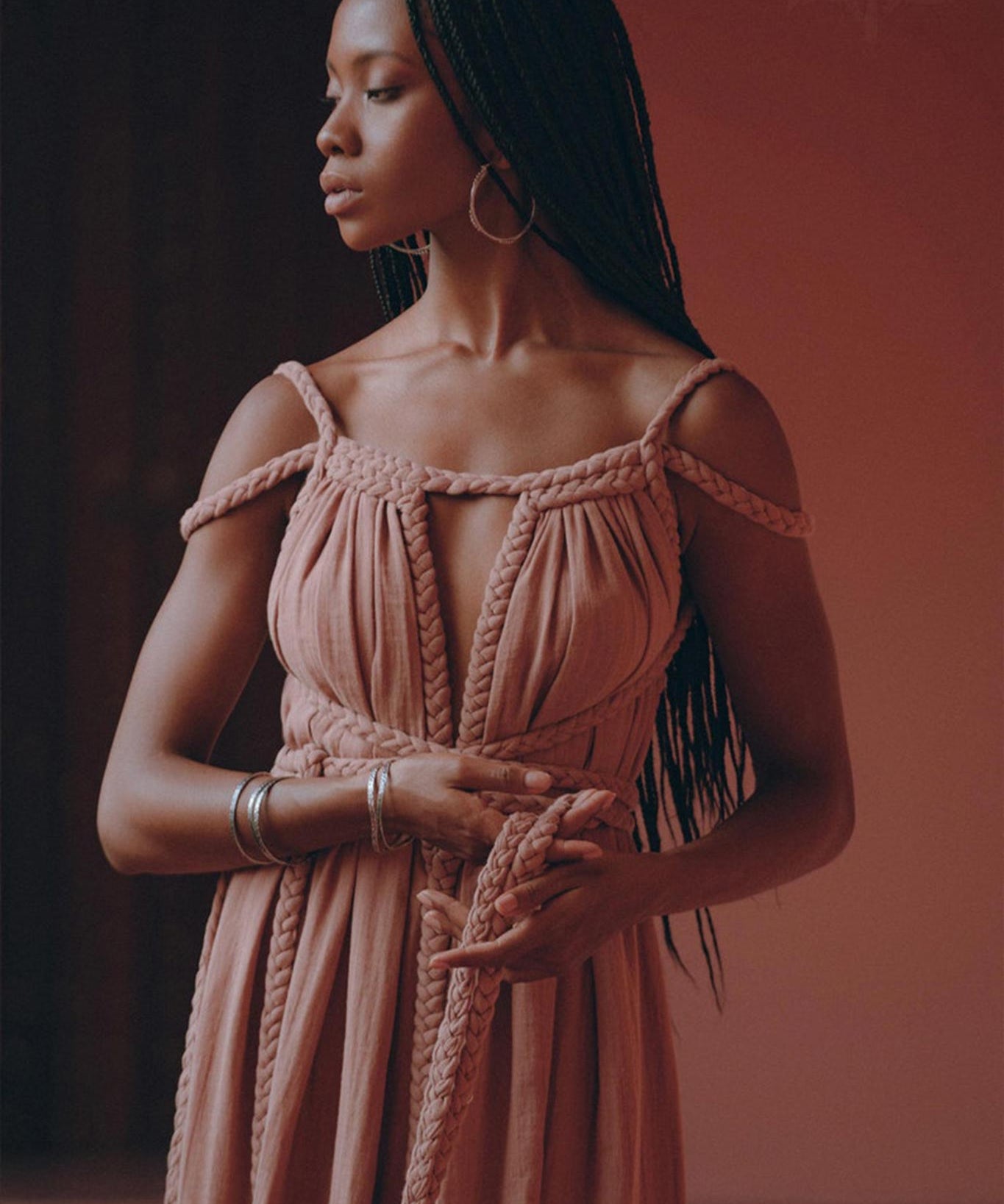 A woman with long braided hair stands gracefully, dressed in a flowy off-shoulder Dusty Pink Greek Goddess Prom Boho Dress by AYA Sacred Wear, complemented by hoop earrings, against a warm, peach-colored background. She appears contemplative as she holds the fabric of her dress in her hands.