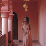 A woman in a Dusty Pink Greek Goddess Prom Boho Dress by AYA Sacred Wear stands in a pink-walled corridor, holding a long necklace. Above her, three ornate round lamps hang from the ceiling, casting a warm glow. A wooden door with an arch is in the background.