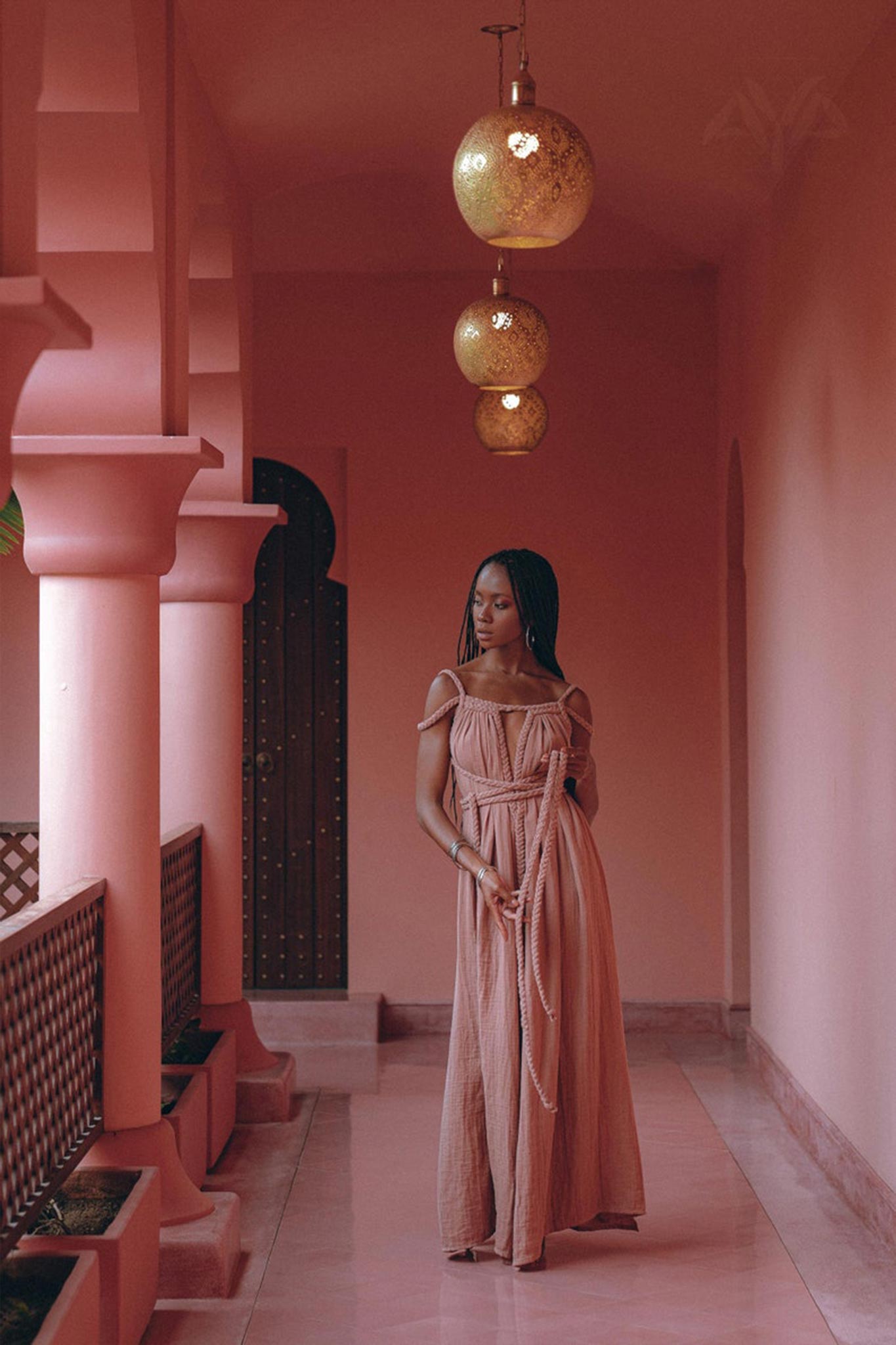 A woman in a Dusty Pink Greek Goddess Prom Boho Dress by AYA Sacred Wear stands in a pink-walled corridor, holding a long necklace. Above her, three ornate round lamps hang from the ceiling, casting a warm glow. A wooden door with an arch is in the background.