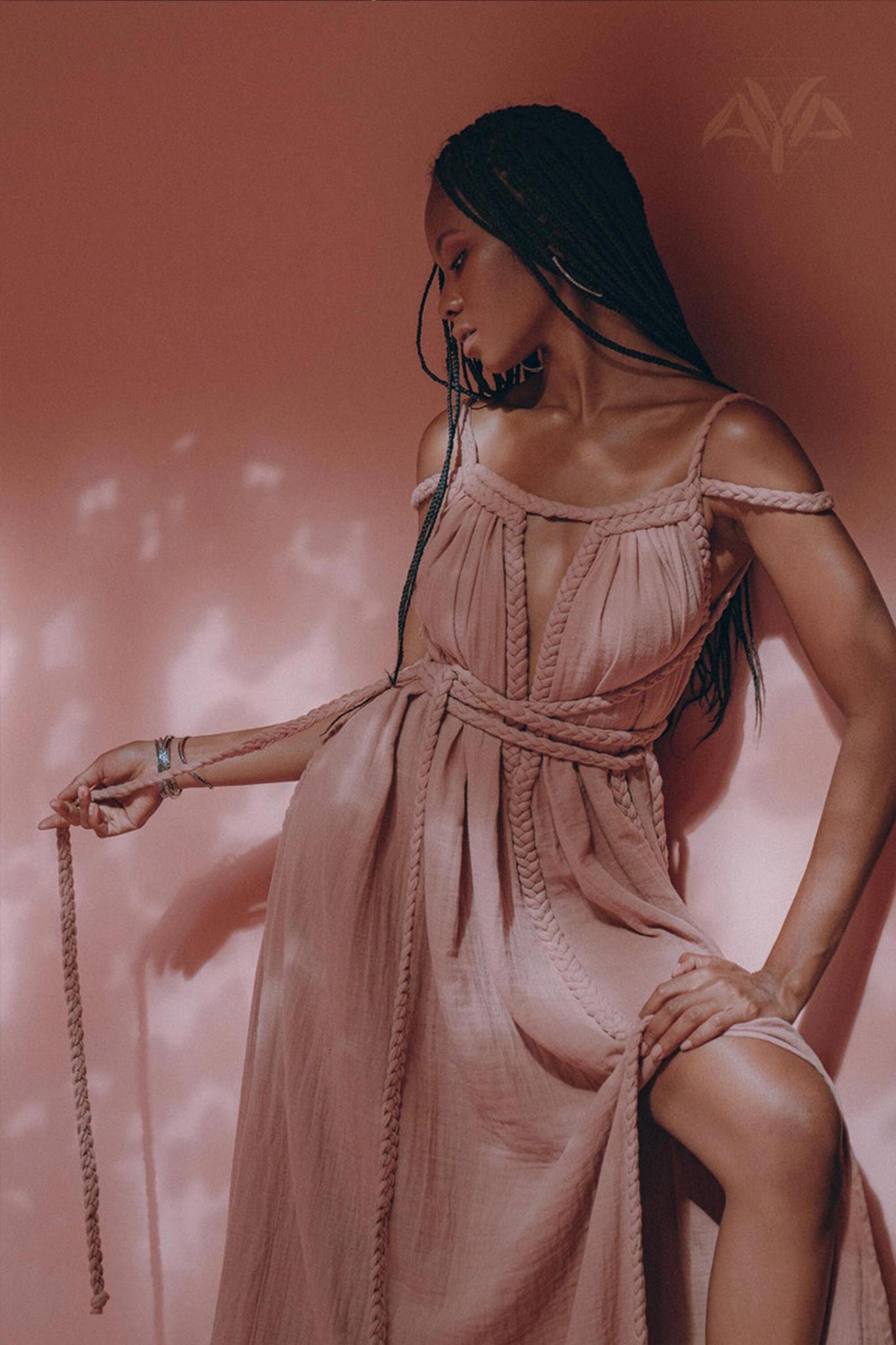 A person wearing the Dusty Pink Greek Goddess Prom Boho Dress from AYA Sacred Wear poses against a soft pink backdrop with shadowed patterns. Crafted from organic cotton, this dress exudes a serene and elegant atmosphere as they gracefully hold a rope.