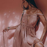 A person wearing the Dusty Pink Greek Goddess Prom Boho Dress from AYA Sacred Wear poses against a soft pink backdrop with shadowed patterns. Crafted from organic cotton, this dress exudes a serene and elegant atmosphere as they gracefully hold a rope.