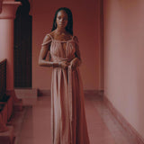 A woman wearing a Dusty Pink Greek Goddess Prom Boho Dress from AYA Sacred Wear stands in a dusty pink corridor adorned with ornate archways and hanging lanterns. The polished floor gleams beneath her, and a wooden bench sits to one side. With a serene expression, she gazes forward, embodying effortless elegance.